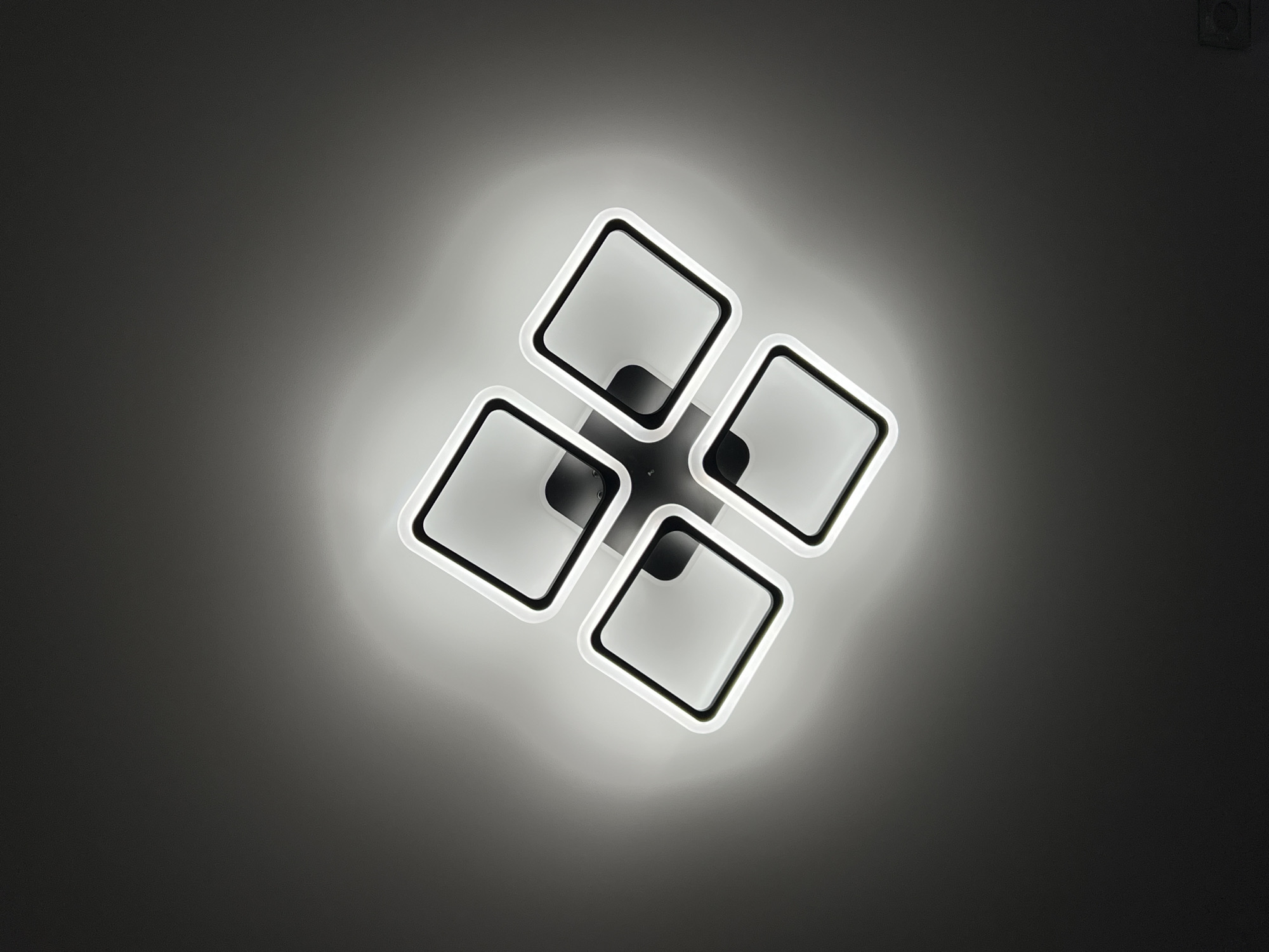 The image presents a close-up view of a modern ceiling light fixture against a gray backdrop. The fixture consists of four square-shaped modules, each featuring a black border with a white light-emitting surface inside. The modules are arranged in a clustered formation with the corners of the squares converging towards the center. The center of the fixture appears to have a black mounting plate. The light emitted from the fixture creates a diffused halo effect on the surrounding gray background. The lighting is bright and uniform across the visible light-emitting surfaces. A small square object is visible on the top right of the image. There is no information in the image to conclude the location is in the city of Chaikovsky, Russia.