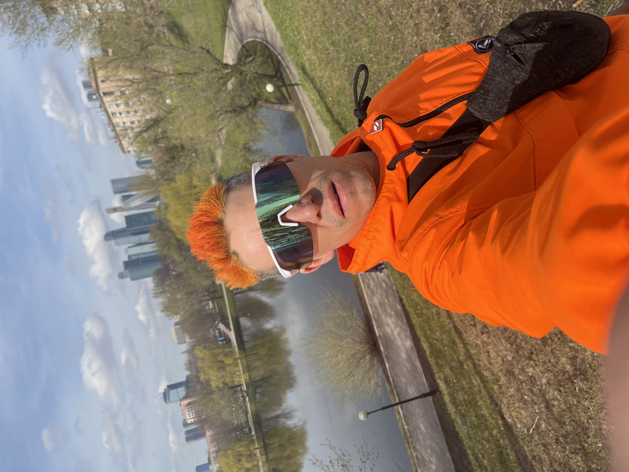 A person with orange hair and sunglasses is taking a selfie outdoors, with an orange jacket draped over their shoulder. In the background, a body of water reflects the sky and buildings. The location appears to be Moscow, Russia, based on the architecture of the buildings in the background. Trees line the edge of the water, and a grassy area is visible alongside a paved path. A lamppost is also visible along the path. The person is wearing a black bag on their shoulder.
