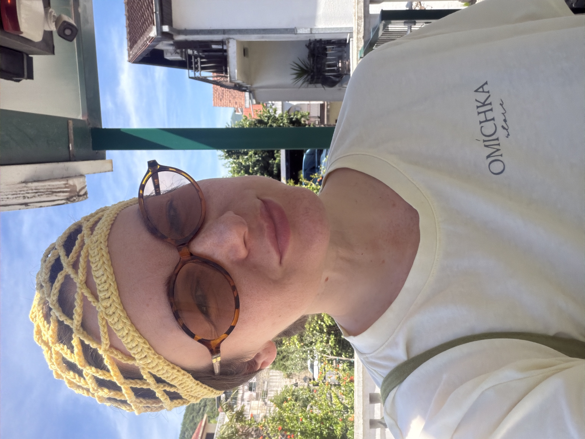 A person wearing sunglasses and a yellow crocheted head covering is shown from a low angle. The person is wearing a light yellow t-shirt with the inscription "OMICHKA slave" in dark gray lettering. They are standing outdoors, under a green awning. In the background, there are buildings and trees, suggesting the location is Meljine, Montenegro.