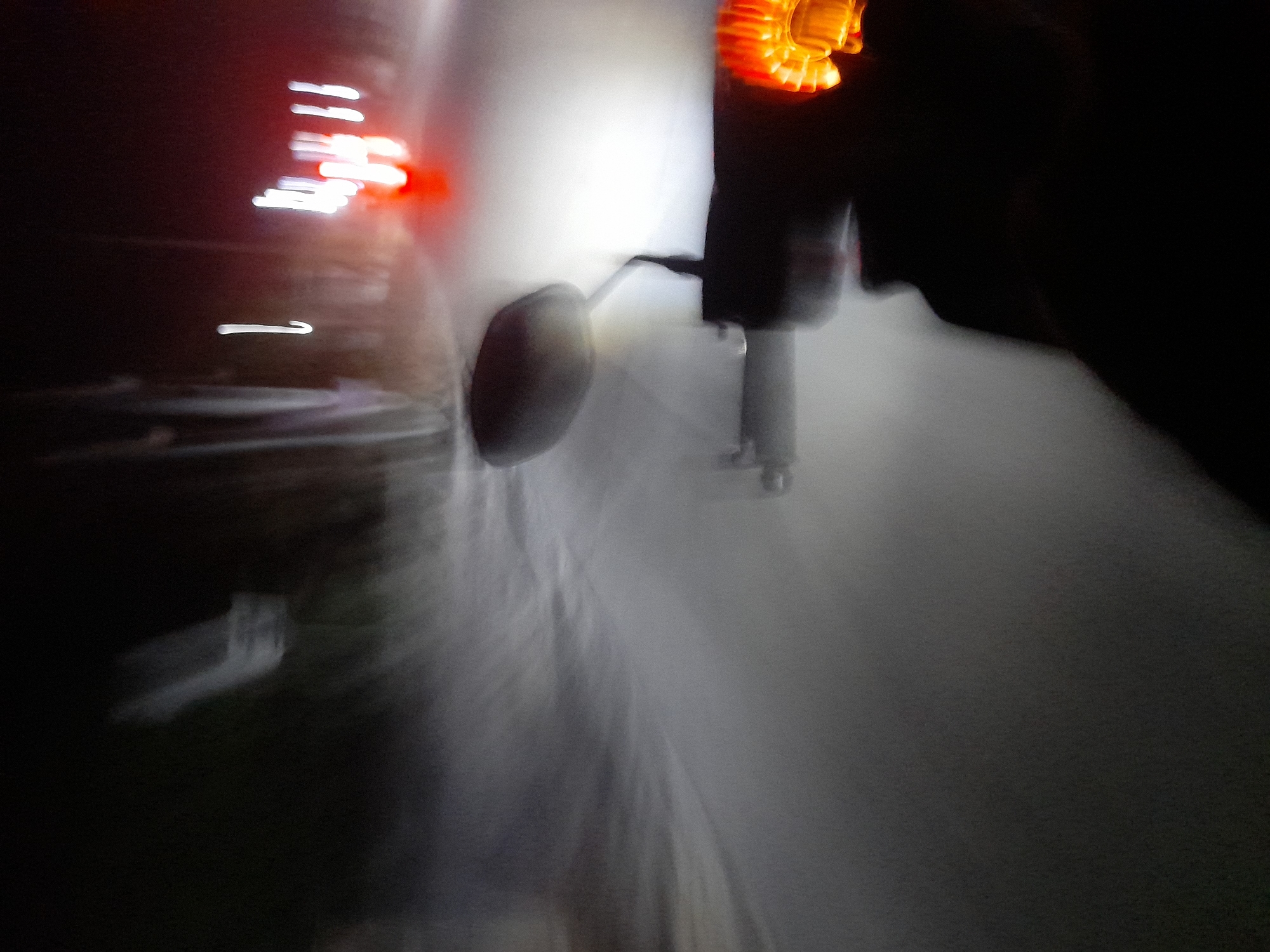 The image depicts a blurry, nighttime scene from the perspective of someone riding a motorcycle in Jetaksari, Indonesia. The road ahead is illuminated by the motorcycle's headlight, creating streaks of light due to motion blur. In the distance, other vehicles with illuminated taillights are visible. The environment appears to be a road, possibly in a rural or suburban area, with some indistinct shapes that could be trees or structures along the side. The overall impression is one of movement and low visibility, characteristic of driving at night in an area with limited street lighting.