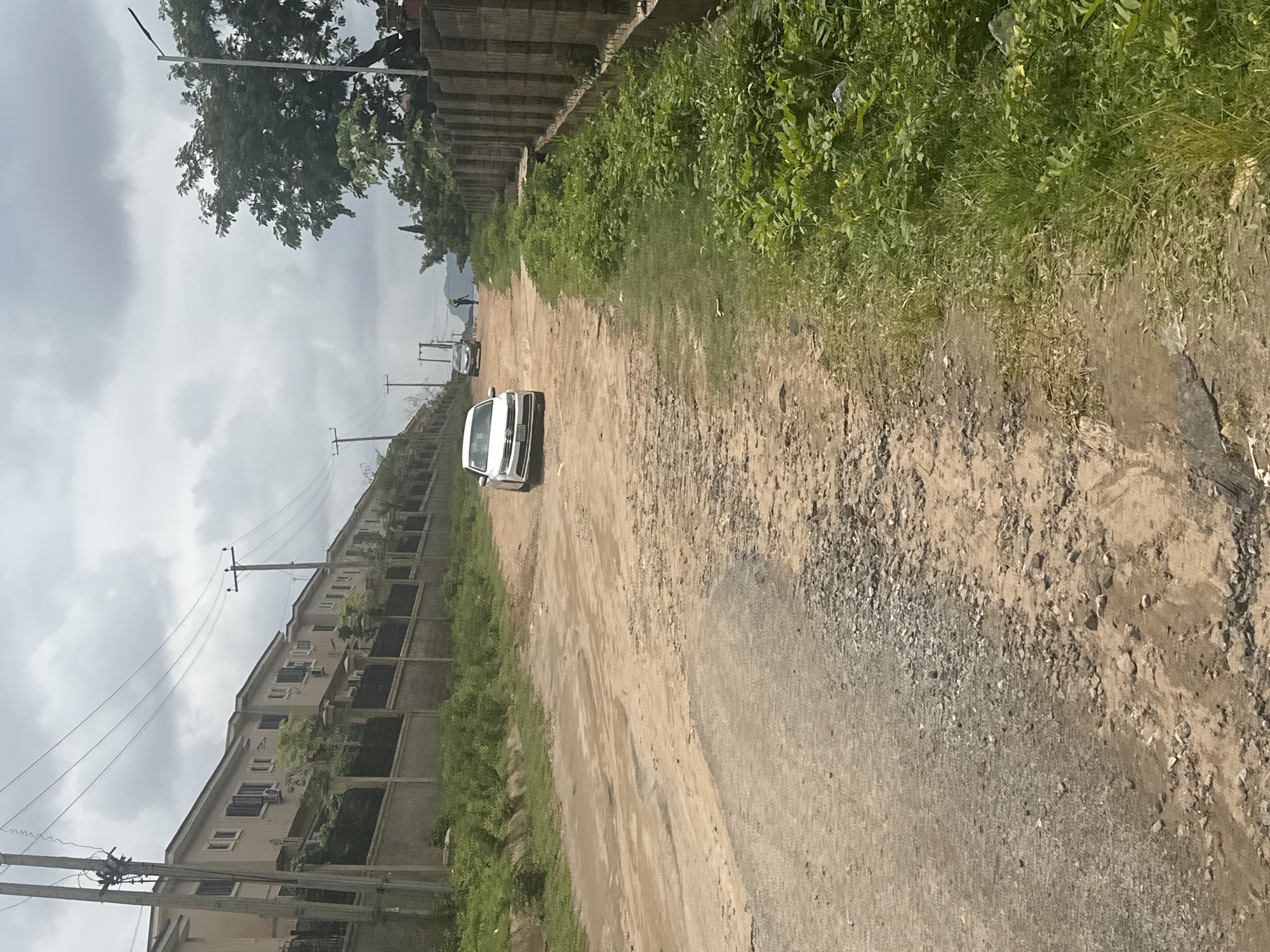 The image depicts a dirt road in Gwarinpa, Nigeria, bordered by a line of residential buildings on the left and a concrete block wall and vegetation on the right. A white sedan is parked on the left side of the road, facing away from the camera. Further down the road, another car is visible, and a person is walking in the distance. The sky is overcast with clouds. The road surface is uneven and appears to be unpaved, with patches of gravel and dirt. Lush green vegetation grows along the sides of the road and behind the wall on the right. The architecture of the houses suggests a modern residential development. The overall scene captures a typical streetscape in a suburban area.