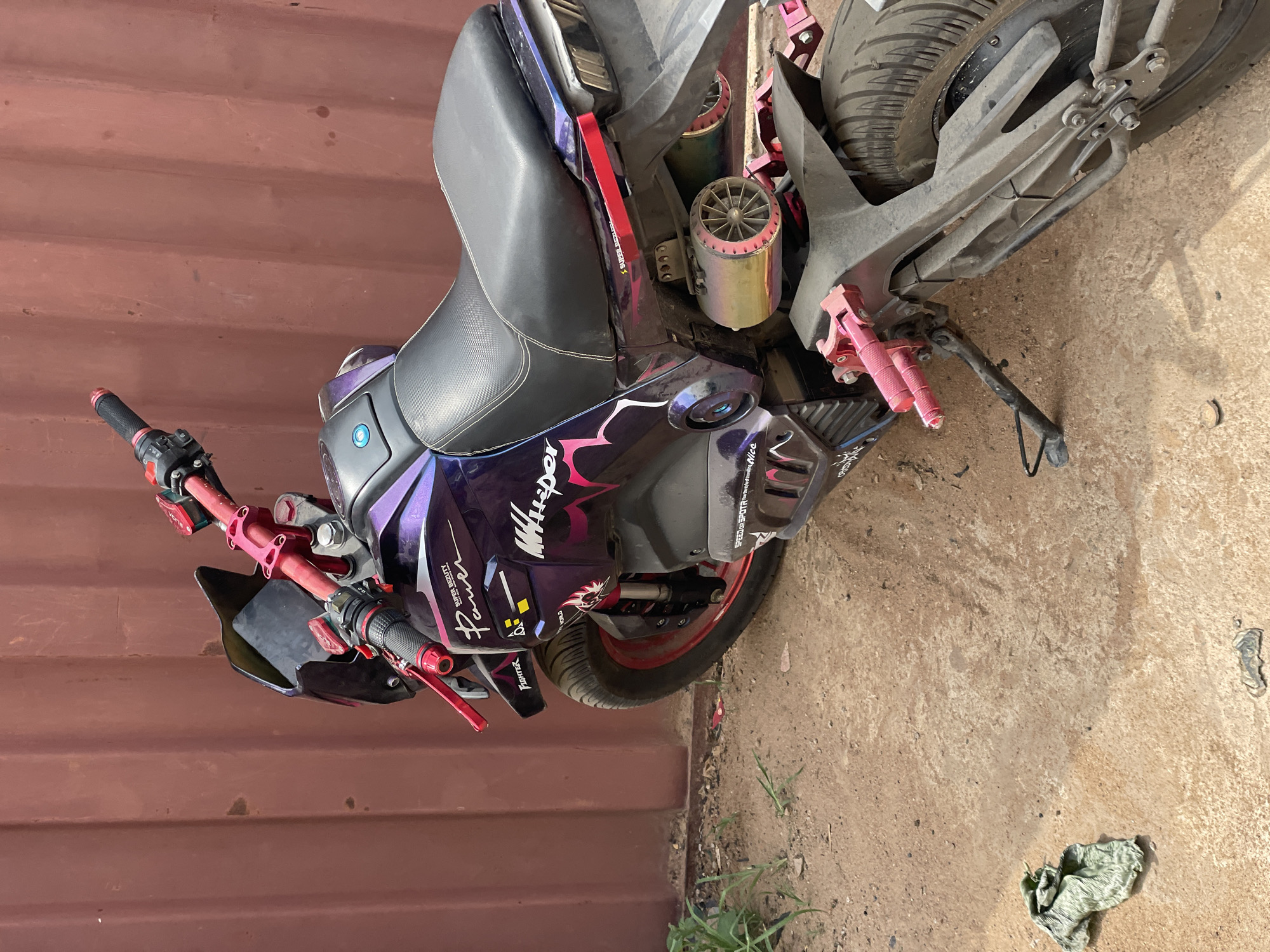 An eye-level, medium shot shows a customized purple and black moped parked on a concrete surface next to a maroon metal wall, possibly a shipping container, in Nkpor, Nigeria. The moped faces towards the left of the frame. The handlebars are red with black grips, and the seat is black. Graphics on the body read "Whisper" and "Fighter." Pink accents appear on the footrests. The rear tire is visibly dirty. The kickstand is deployed. A green cloth or rag rests on the ground near the bottom right of the frame. Vegetation is sparse along the base of the concrete.