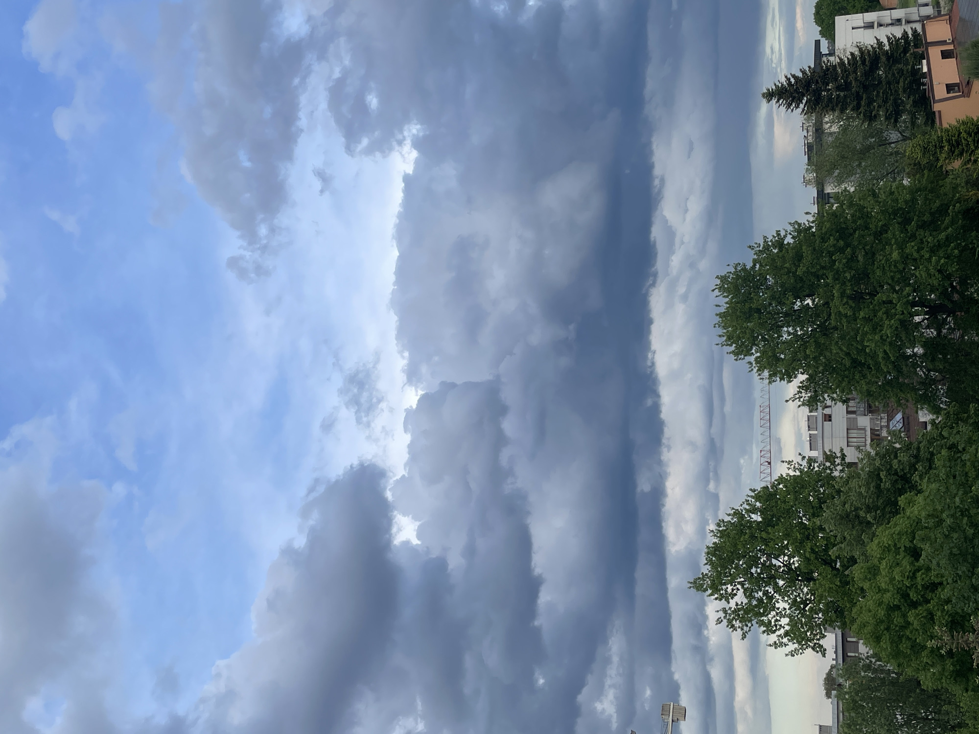 The image shows a view of the sky above a city. A mix of blue sky and ominous, dark gray clouds dominate the frame. The clouds appear in varying formations, some puffy and scattered, others forming a more solid, linear band. Parts of buildings and trees are visible along the right edge. In the distance, a construction crane can be spotted. The overall impression is of an approaching storm.