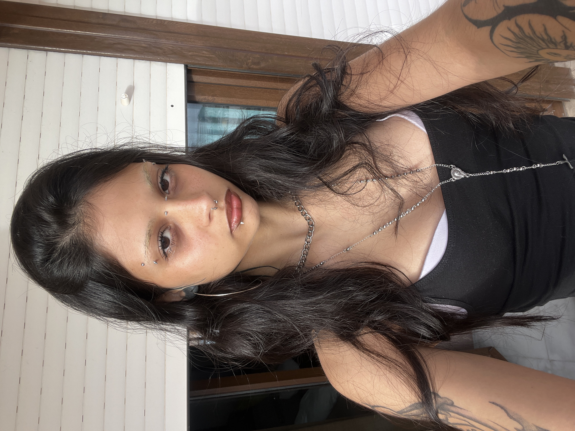 A young woman is taking a selfie indoors. She has dark hair, light skin, and multiple facial piercings. She is wearing a black tank top and silver necklaces. Tattoos are visible on her arms. The background shows blinds and a window with a dark frame. The location is Arroyo De La Miel-Benalmadena Costa, Spain, inferred from the architectural details in the background.