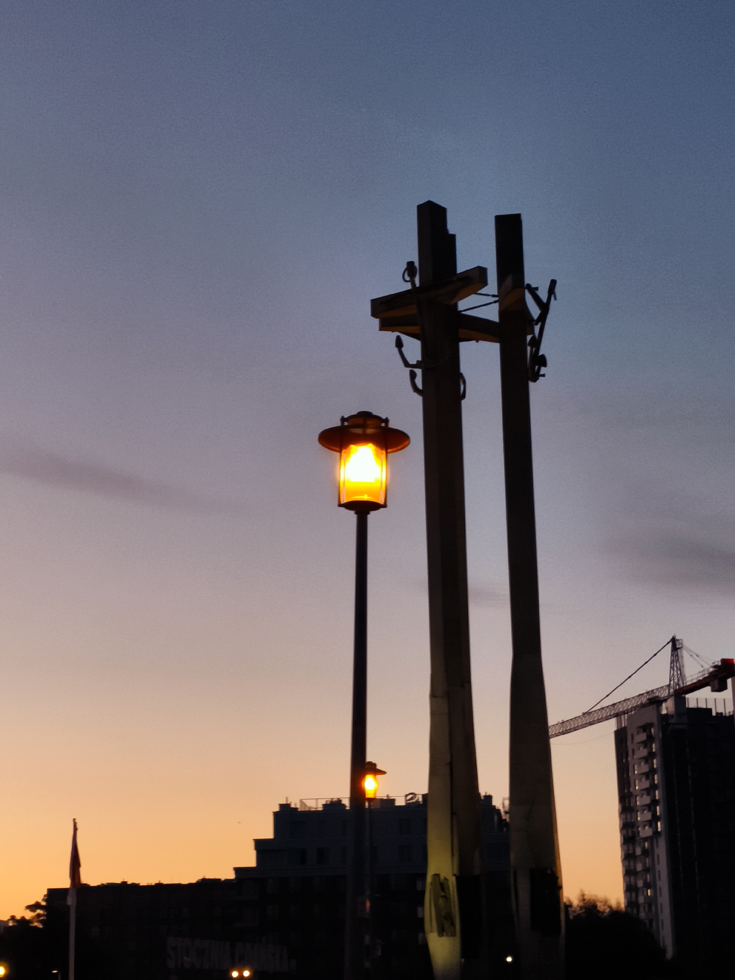 The image depicts a twilight scene in Gdańsk, Poland, likely near the Shipyards. In the foreground, a street lamp with an orange glow stands tall. Behind it, a monument consisting of three large concrete pillars is visible, with two of them framing a third, narrower pillar. The monument is silhouetted against the dusky sky. To the right, a modern high-rise building under construction is partially visible, with a construction crane positioned above it. In the background, silhouetted buildings and trees line the horizon. A red flag flies on a flagpole to the left, and text, possibly "STOCZNIA GDAŃSKA" (Gdańsk Shipyard), is faintly legible on a dark structure in the lower portion of the image. The sky transitions from a pale orange near the horizon to a deep blue at the top, with some wispy clouds.