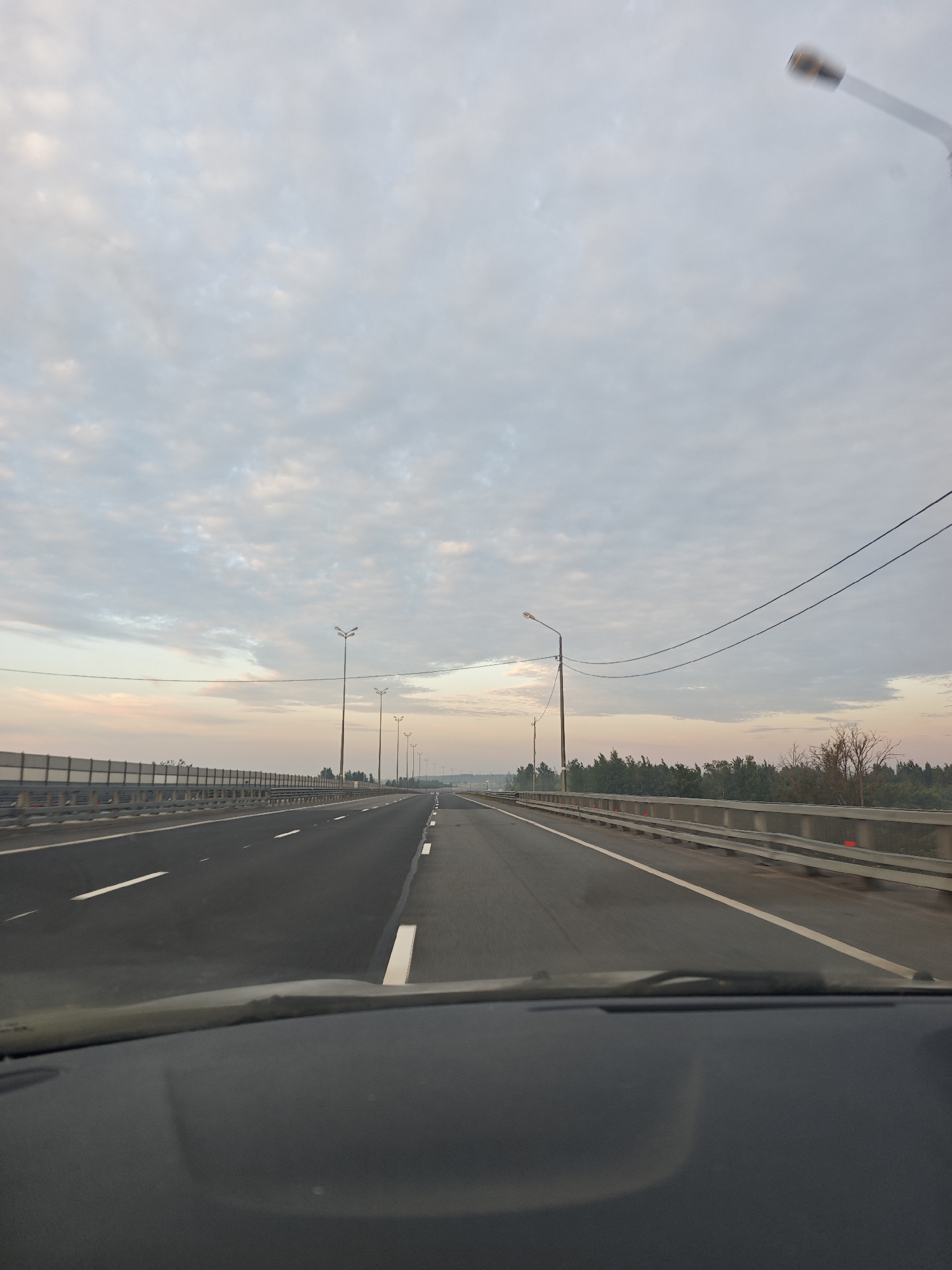The image shows a view from a vehicle driving on a highway in Торфяное, Russia, likely during dawn or dusk, as indicated by the soft lighting and cloudy sky. The highway appears to be a multi-lane road with clear lane markings. On the left side of the highway, there is a sound barrier and a row of streetlights. On the right side, there is a concrete barrier and trees lining the edge of the road. The sky is filled with scattered clouds, some tinged with pink and orange hues. The dashboard of the vehicle is visible in the foreground, partially obscuring the lower part of the image. There are no people visible in the scene, and the focus is on the road and the surrounding landscape. The overall impression is one of a peaceful, solitary journey on an open road.