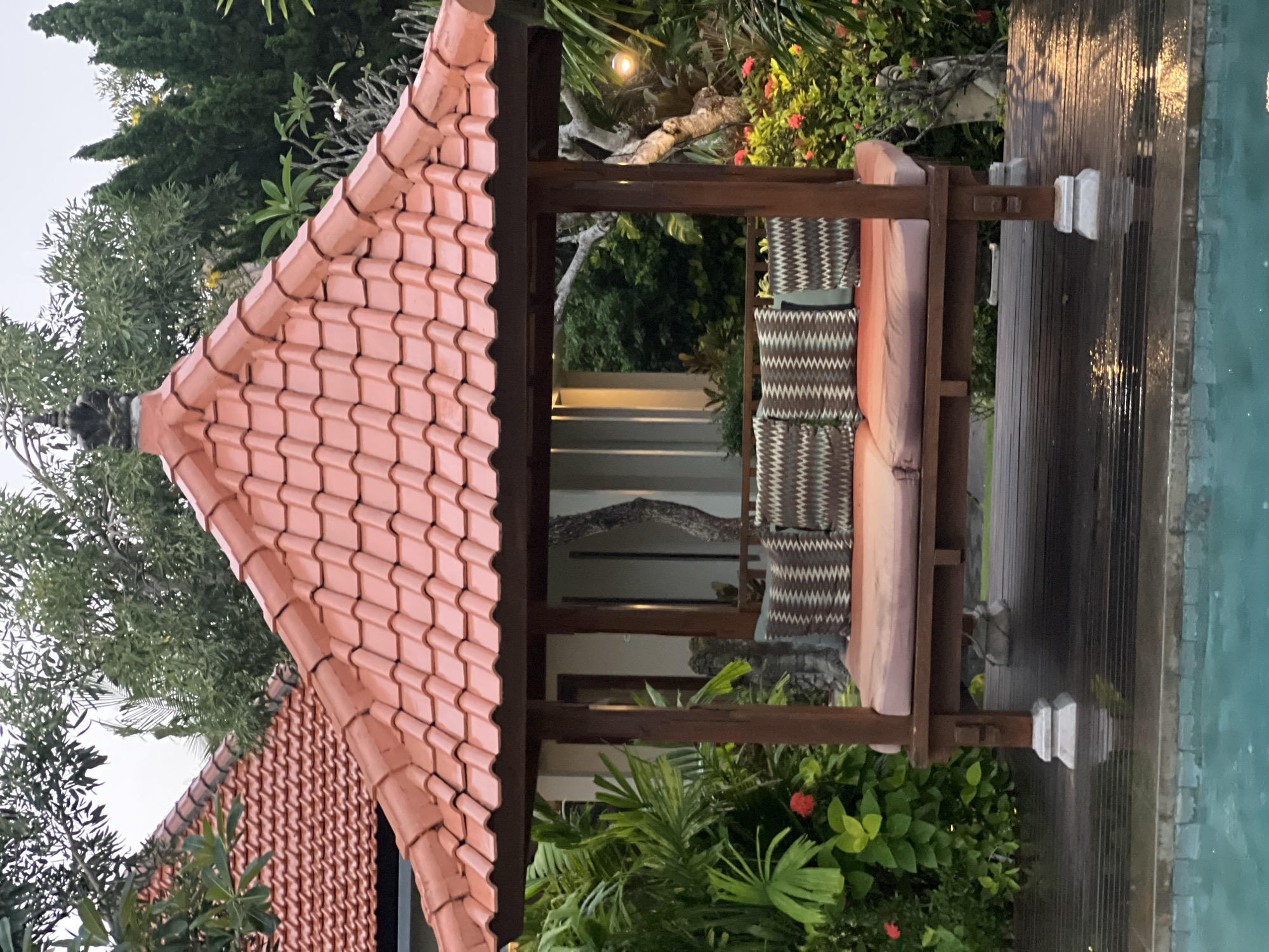 The image depicts a small gazebo or pavilion with a red-tiled roof in an outdoor setting, likely a garden or resort area in Kuta Selatan, Indonesia. The gazebo features dark wooden posts and beams, with a built-in bench or daybed arrangement that includes cushions with a patterned design. Lush tropical vegetation surrounds the gazebo, and a body of water (possibly a pool) is visible on the right side of the frame. In the background, a structure or building with a light-colored facade is partially visible through the gazebo's open sides. The light suggests either early morning or late afternoon. No people are visible in the image.