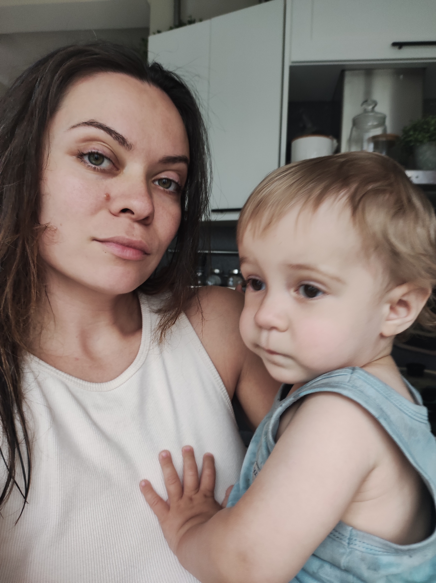 A woman and a young child are posing for a selfie indoors, likely in a kitchen. The woman has dark brown hair, green eyes, and fair skin, and is wearing a white tank top. She has a neutral expression and is looking directly at the camera. The child, with blonde hair, is wearing a blue tank top. The child has a pensive expression and is looking to the side. The child's hand is resting on the woman's chest. Behind them, a kitchen setting is visible, including white cabinets and various kitchen items. The specific location in Chaikovsky, Russia, cannot be determined from the image alone.