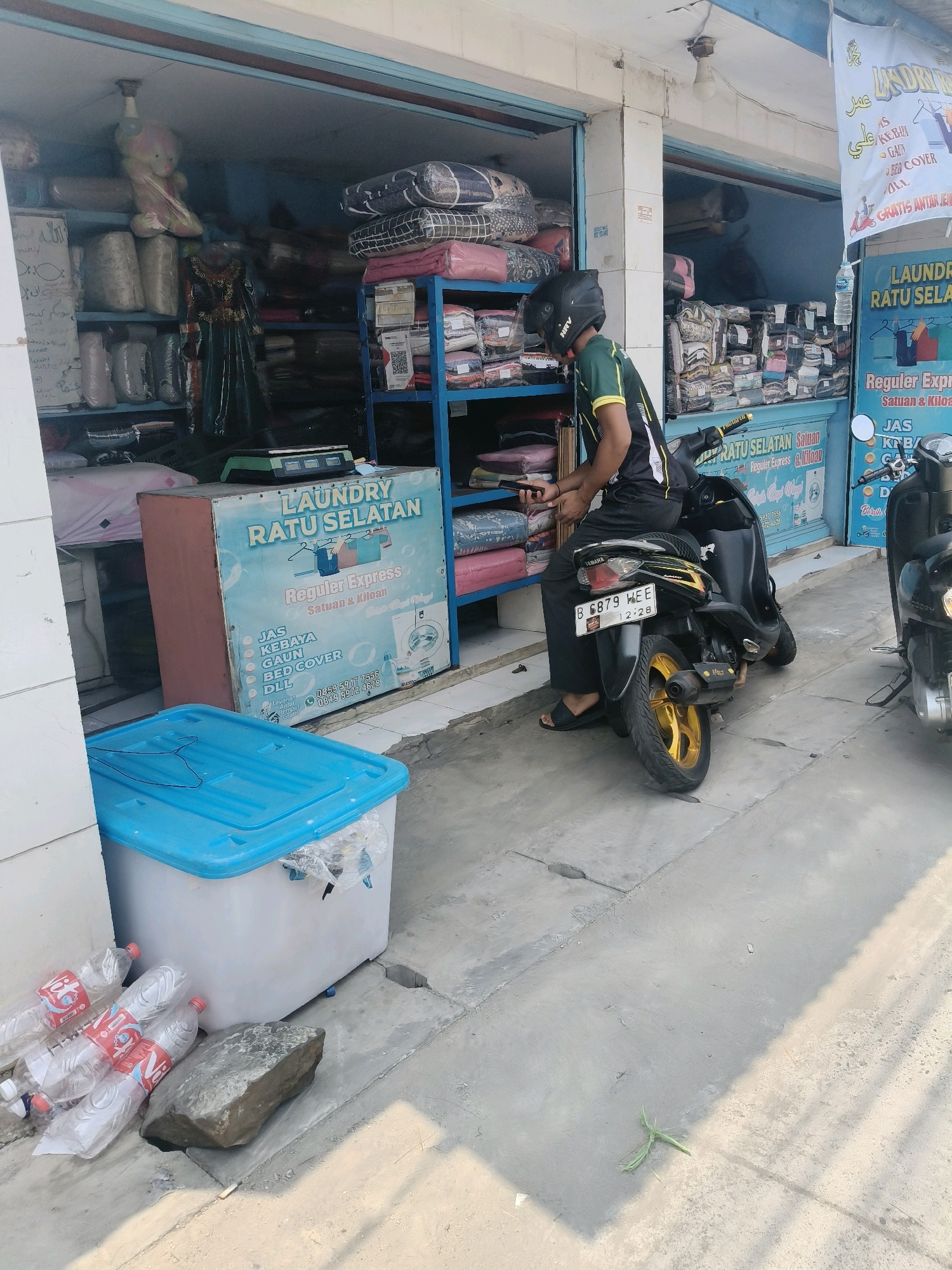 The image depicts a street-level view of a laundry service shop in South Tangerang, Indonesia. The shop's exterior features shelves filled with neatly folded laundry, some still in plastic packaging. A man wearing a helmet and a green and black shirt is seated on a black motorcycle parked in front of the shop. He is holding a smartphone in his right hand. The motorcycle has a license plate displaying "B 6879 WEE". Adjacent to the shop, a large blue-lidded plastic container sits on the pavement, with several empty plastic water bottles and a large rock nearby. The pavement is made of concrete, and a drain grate is visible. A second, darker motorcycle is parked further down the street to the right. The scene is illuminated by natural sunlight, casting shadows on the ground. The overall environment suggests a commercial area on a sunny day.