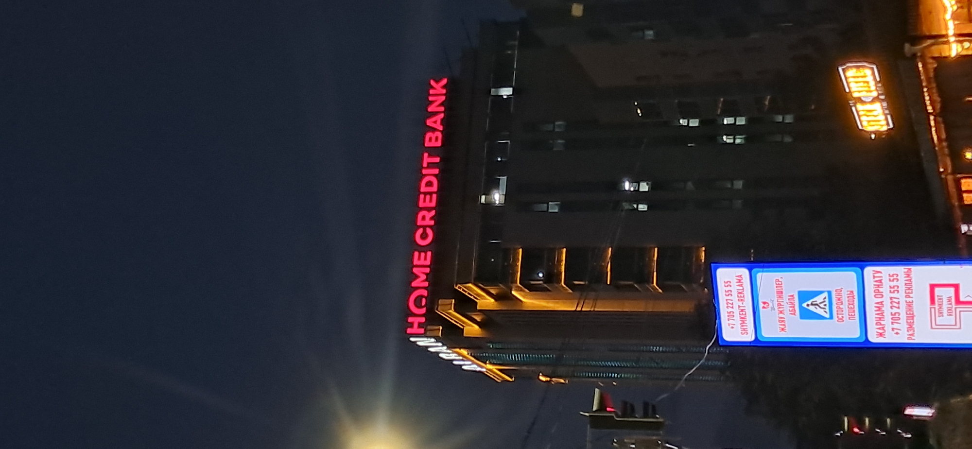 This is a nighttime, eye-level, low-angle shot of a building facade with illuminated signage. The primary subject is a tall, modern building with numerous windows, many of which are lit from within. A large, bright red neon sign vertically displays "HOME CREDIT BANK" on the left side of the building. To the right of this, a horizontal illuminated sign with a blue border contains text and symbols in both Russian and what appears to be Kazakh. The text includes contact information, "SHYMKENT-REKLAMA," and warnings such as "ОСТОРОЖНО, ПЕШЕХОДЫ" (Caution, Pedestrians) with a pedestrian crossing symbol. There's also an indication for "ЖАРНАМА ОРНАТУ" (Install Advertisement) and "РАЗМЕЩЕНИЕ РЕКЛАМЫ" (Ad Placement). The overall scene suggests an urban environment at night, possibly a commercial or financial district, given the bank signage and advertising. The lighting is artificial, with the neon and illuminated signs creating the main light sources. The dark sky and the lit windows imply it is well after sunset.
