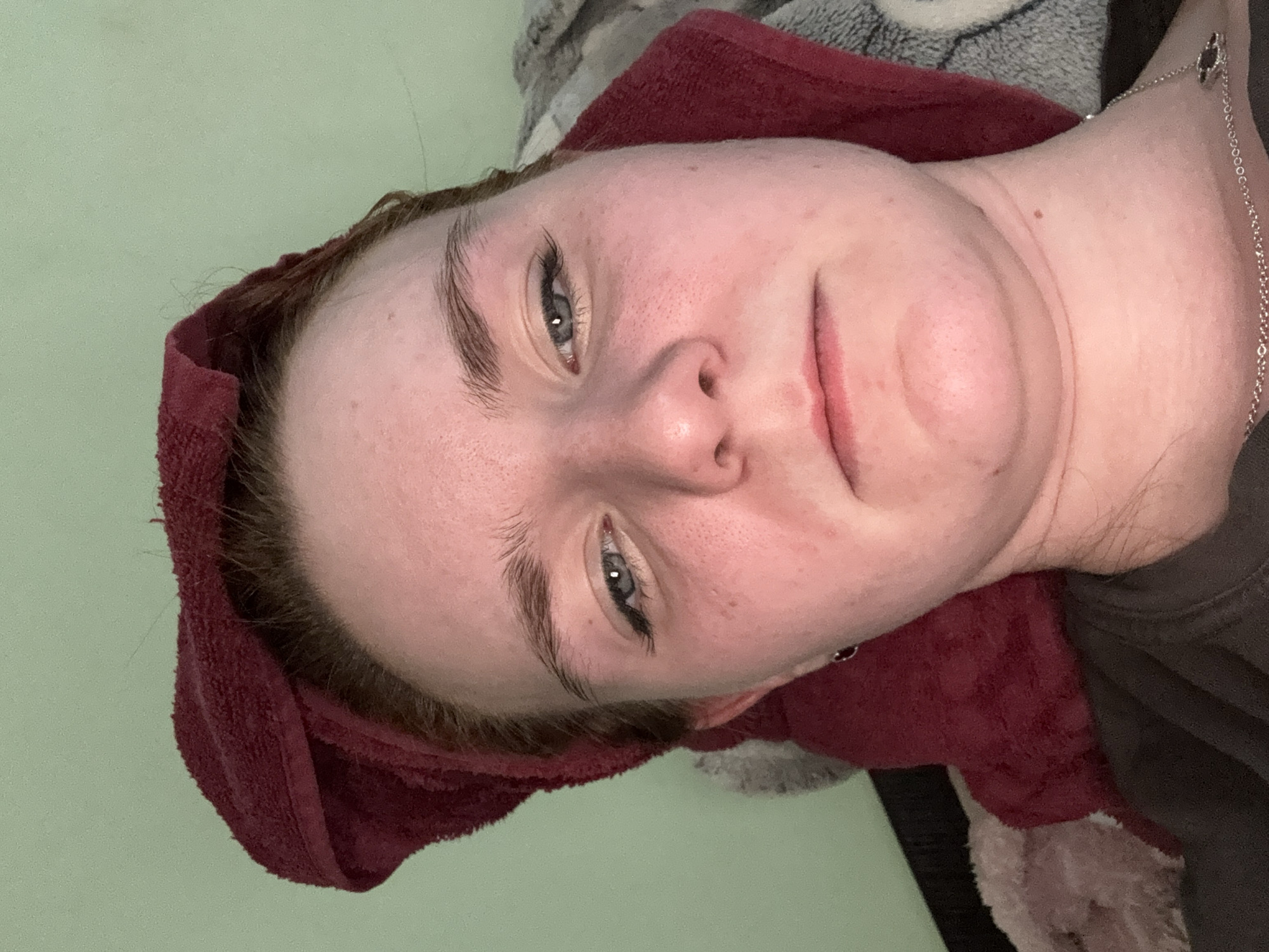 A person with fair skin, blue eyes, and auburn hair is facing the camera. A maroon towel is wrapped around their head. The person appears to be lying down. The background is a light green wall, with a gray and white patterned object behind their head. A silver chain is visible around their neck. The location is Unknown, Russia.