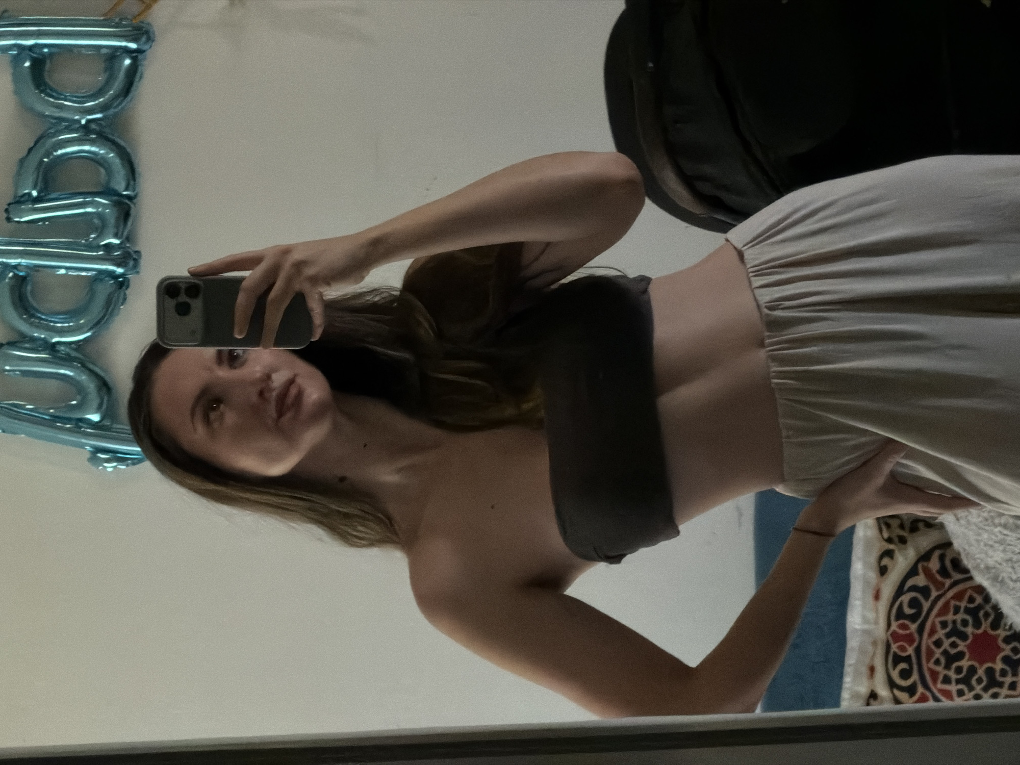 A young woman is taking a mirror selfie in what appears to be a bedroom. She is wearing a dark strapless top and light-colored wide-leg pants. She holds a smartphone in her right hand, capturing her reflection. Behind her, a wall is visible with a string of light blue "baby" balloons. The room has a muted color palette, with a bed or couch visible in the lower left corner with patterned bedding. The lighting is soft, suggesting an indoor setting. There is no indication of the location being Sharm El Sheikh, Egypt, or any other specific city.