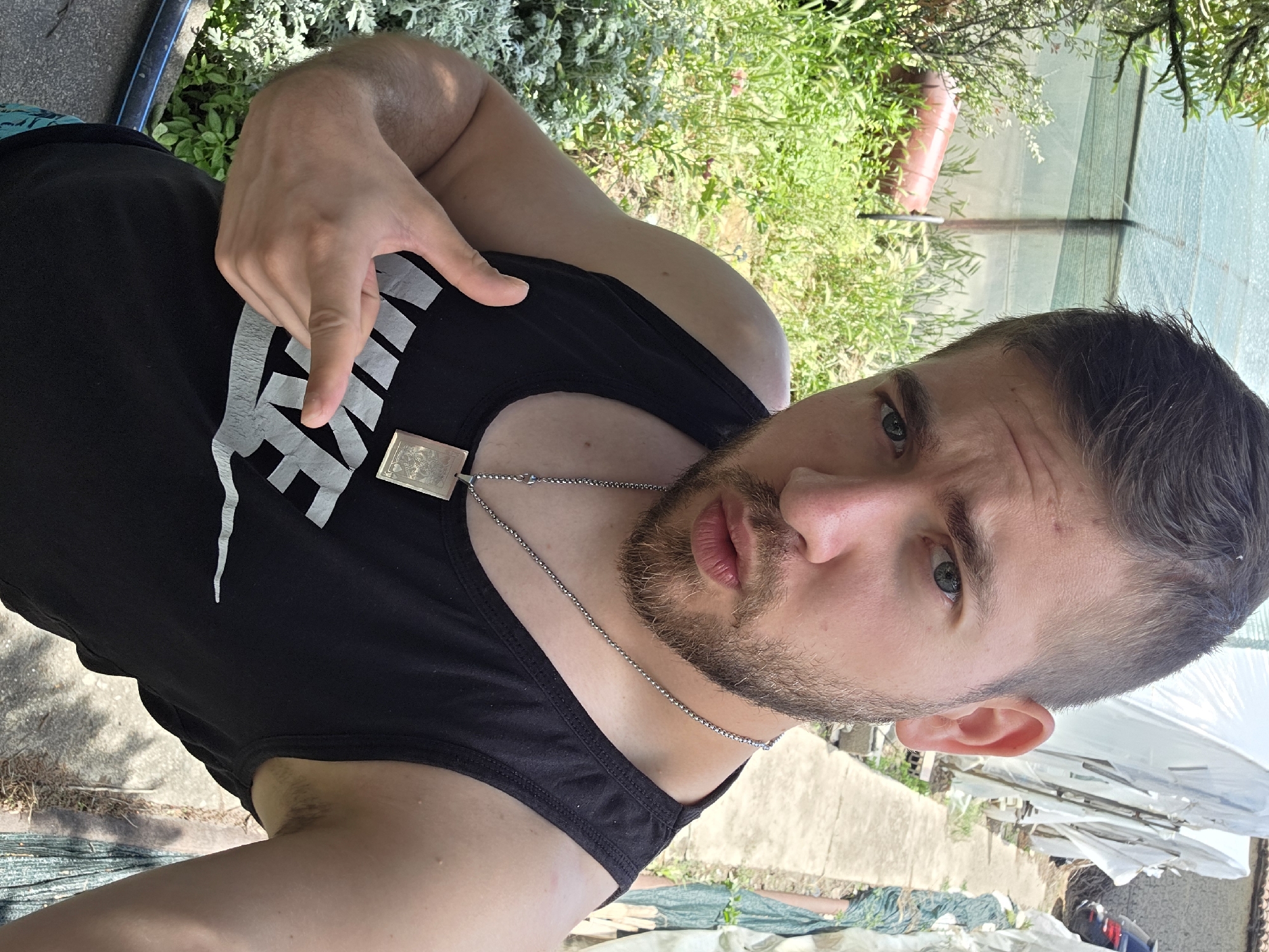 A man with short, dark hair and a beard is wearing a black tank top with white lettering, and a silver chain with a pendant. He is making a gesture with his left hand. The background includes greenery and what appears to be a partial view of a building or structure covered in white material. The location is Yagodovo, Bulgaria.
