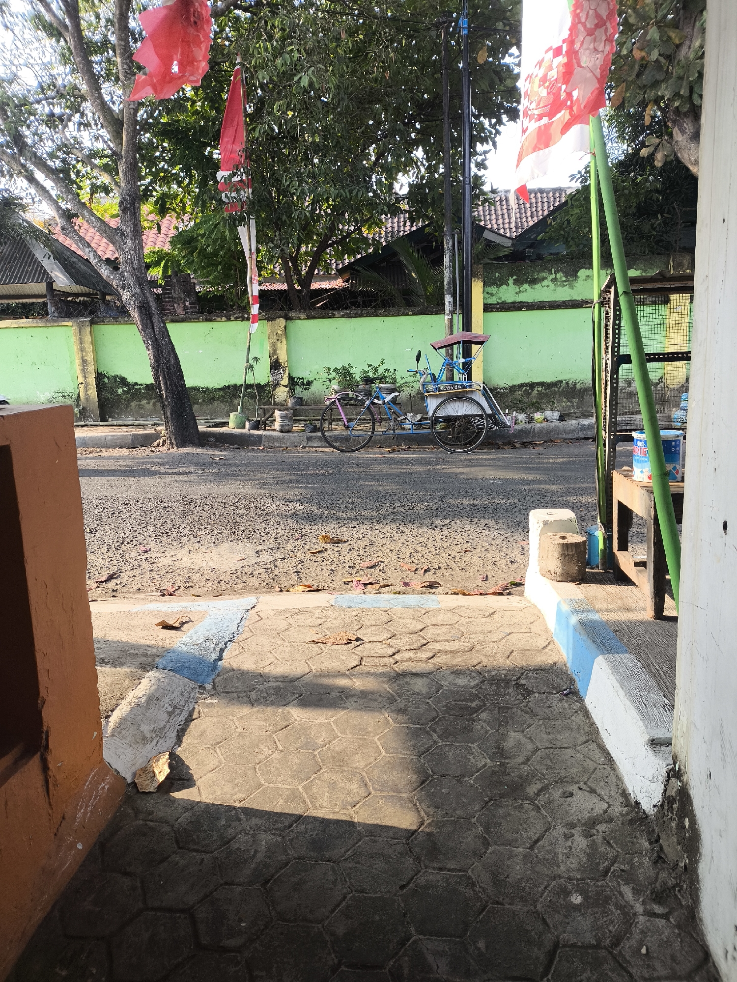 The image depicts a street scene in Citrodiwangsan, Indonesia, bathed in the warm light of late afternoon or early morning. In the foreground, a textured concrete pathway with a hexagonal pattern leads towards the street. The pathway is bordered by a curb painted with blue and white stripes.

The street itself is paved with asphalt and shows signs of wear, with scattered leaves and small stones. To the left, a weathered orange building structure is partially visible. Further down the street, a light green wall runs along the property line, topped with a black metal fence. Behind the wall, houses with red tiled roofs are visible.

A tree with a thick trunk stands prominently near the wall, its branches reaching upwards. A red tattered flag hangs from one of the branches, and another Indonesian flag (red and white) is attached to a bamboo pole nearby.

In the middle ground, a bicycle with a blue frame and a red canopy is parked on the side of the road, appearing to be part of a pedicab or becak. There are no people visible in the frame.

To the right, a tall green pole supports wires, and a rustic wooden table or bench sits adjacent to it. On this surface, there are two cylindrical containers, one blue and white, and another partially obscured. The overall impression is a quiet street corner in a residential or commercial area of Indonesia, likely a warm climate given the lush green foliage.