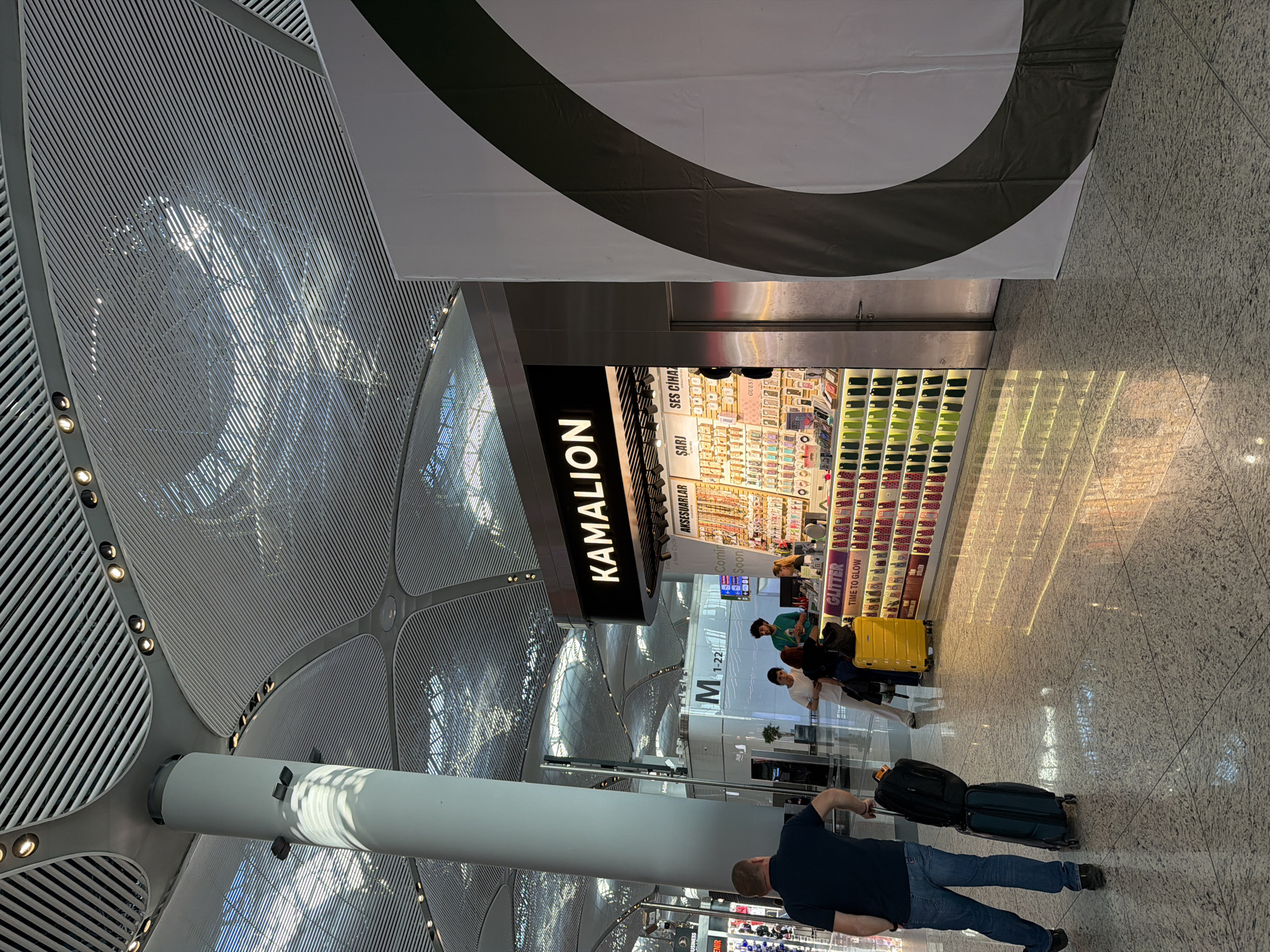 The image depicts an interior view of a modern airport terminal in Arnavutköy, Turkey. A man in a dark t-shirt and jeans is seen from behind, pulling two suitcases, one black and one dark green, across a polished granite floor that reflects the overhead lighting and the storefronts. To his right is a brightly lit kiosk named "KAMALION" which displays an array of accessories, including phone cases, jewelry, and electronic gadgets. Behind the kiosk, a few individuals, one wearing a green shirt, are visible interacting. The terminal features a distinctive architectural design with a high, slatted ceiling supported by large, cylindrical columns. A large, partially visible white banner with a dark curved stripe is positioned to the right of the kiosk, suggesting an advertisement or display. The overall environment suggests a bustling commercial area within an airport.