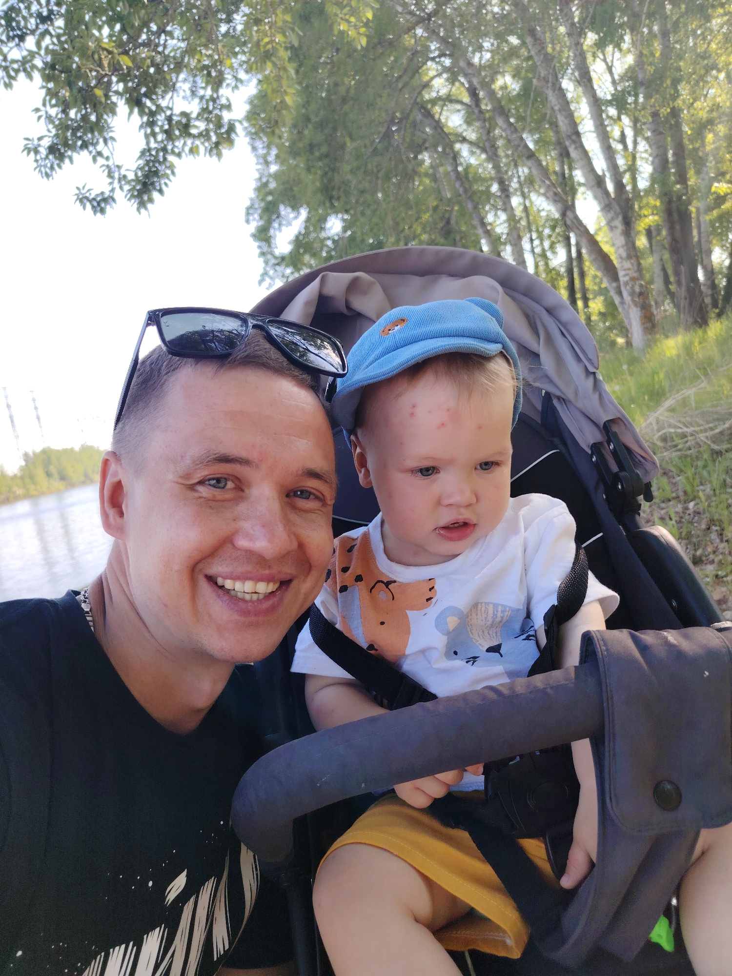 The image captures a selfie of a man and a child in a stroller. The man, on the left, is fair-skinned with short hair and sunglasses perched on his head. He is wearing a black t-shirt with a white design. He is smiling broadly, revealing his teeth. The child, seated in a grey stroller, has fair skin and is wearing a light blue cap with a bear emblem, a white t-shirt with animal prints, and yellow shorts. The child is strapped into the stroller with black safety straps. The background reveals a body of water on the left and lush green trees on the right, suggesting an outdoor setting, possibly a park or riverside area. The location is identified as the city of Chaikovsky, Russia.