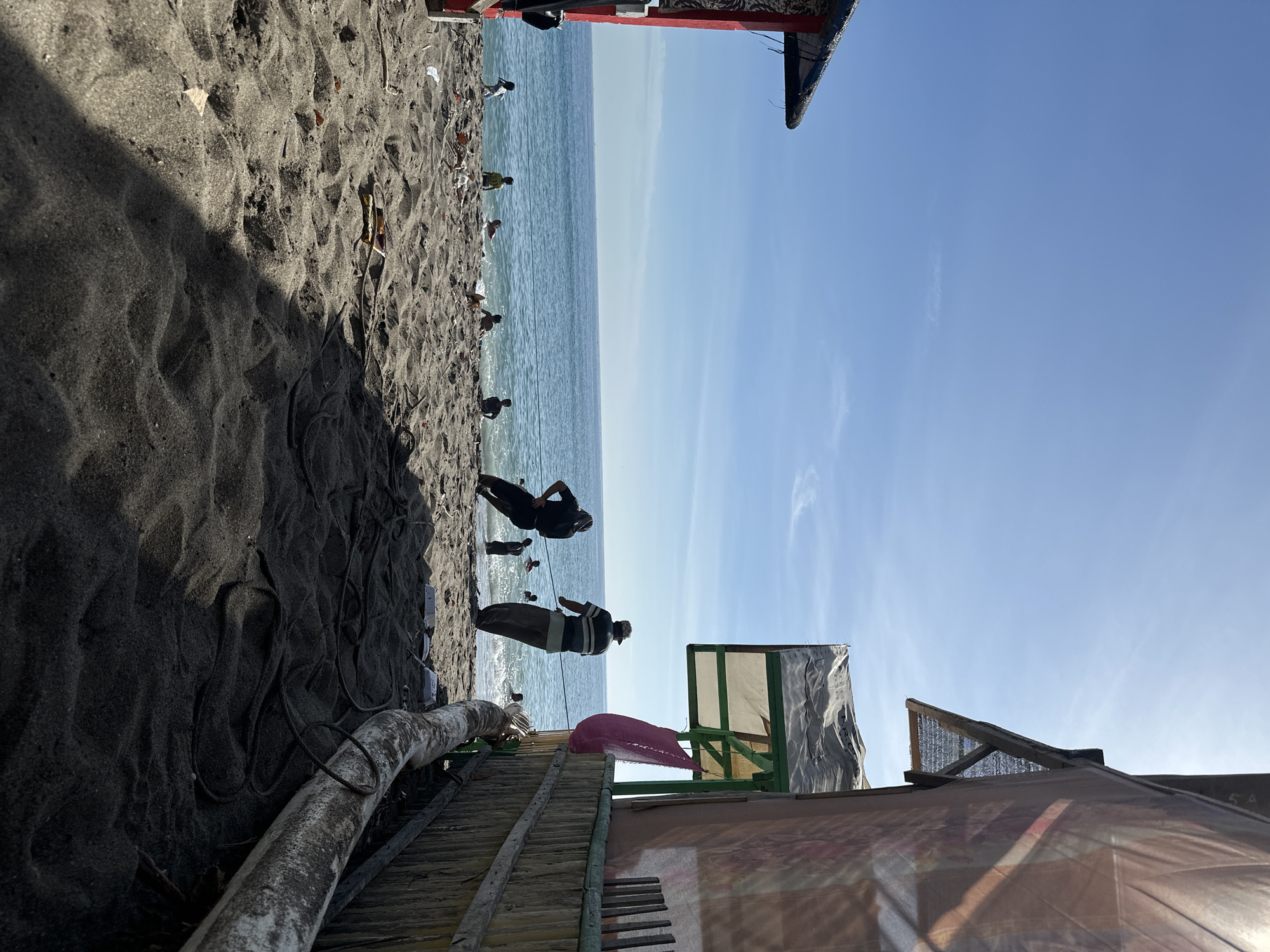 The image depicts a beach scene in Lhokseumawe, Indonesia. The beach has dark sand and is littered with debris. Several people are present, some in the water and others standing on the shore. A makeshift wooden structure or hut is visible in the foreground. The sky is clear blue, and the sea stretches to the horizon. Ropes and logs lie on the beach.