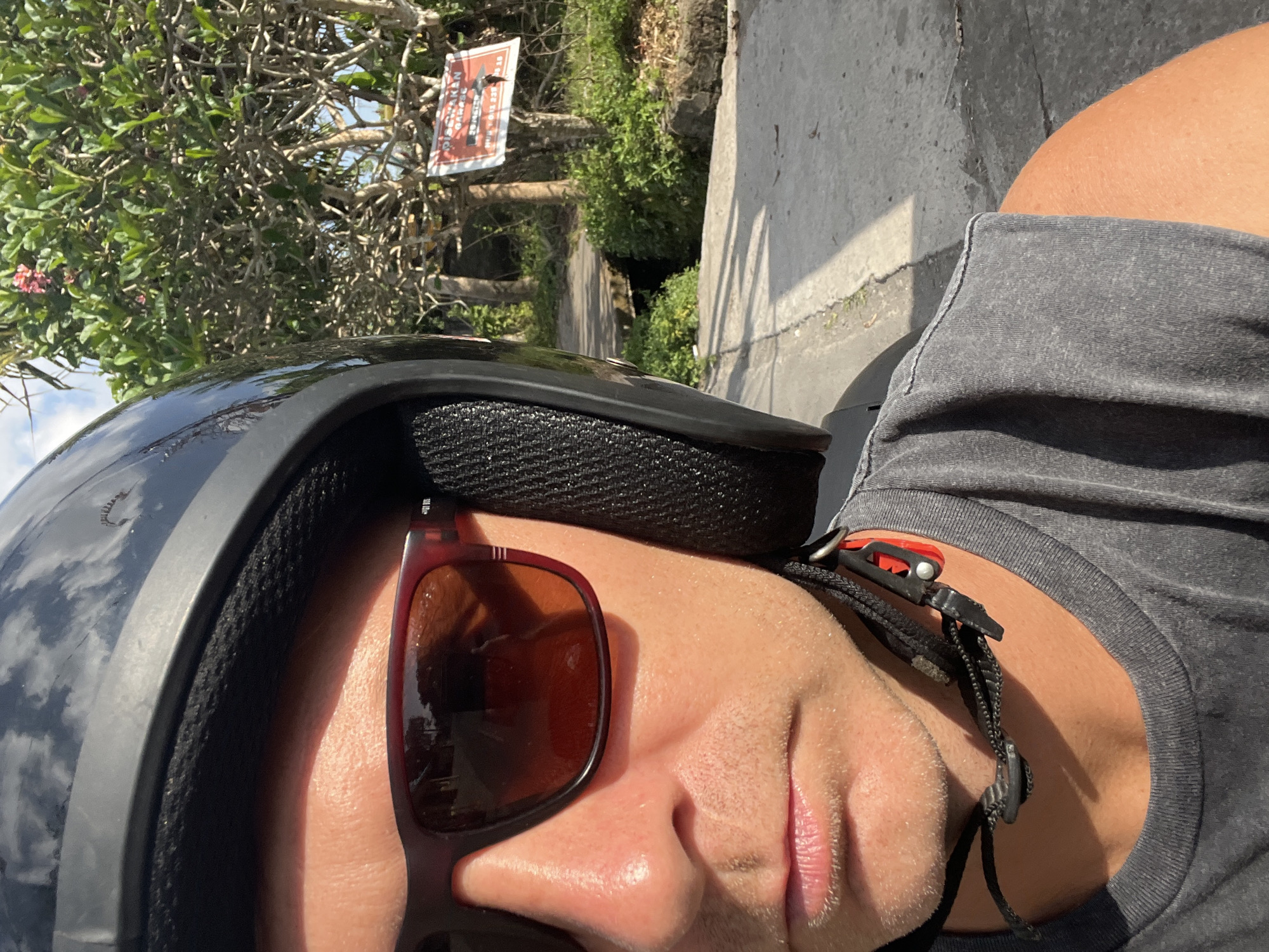 A selfie taken from the perspective of a person wearing a black helmet and sunglasses, seated on a vehicle in Sukawati, Indonesia. The person's face, from the sunglasses down to the neck, is visible, along with a portion of their grey t-shirt. The background features lush green foliage, trees, a paved path, and a sign. The sign appears to be a warning, with text that includes "PERINGATAN GARAGE" and a phone number. The sky is visible at the top of the image, reflecting on the helmet. The lighting suggests it is daytime.