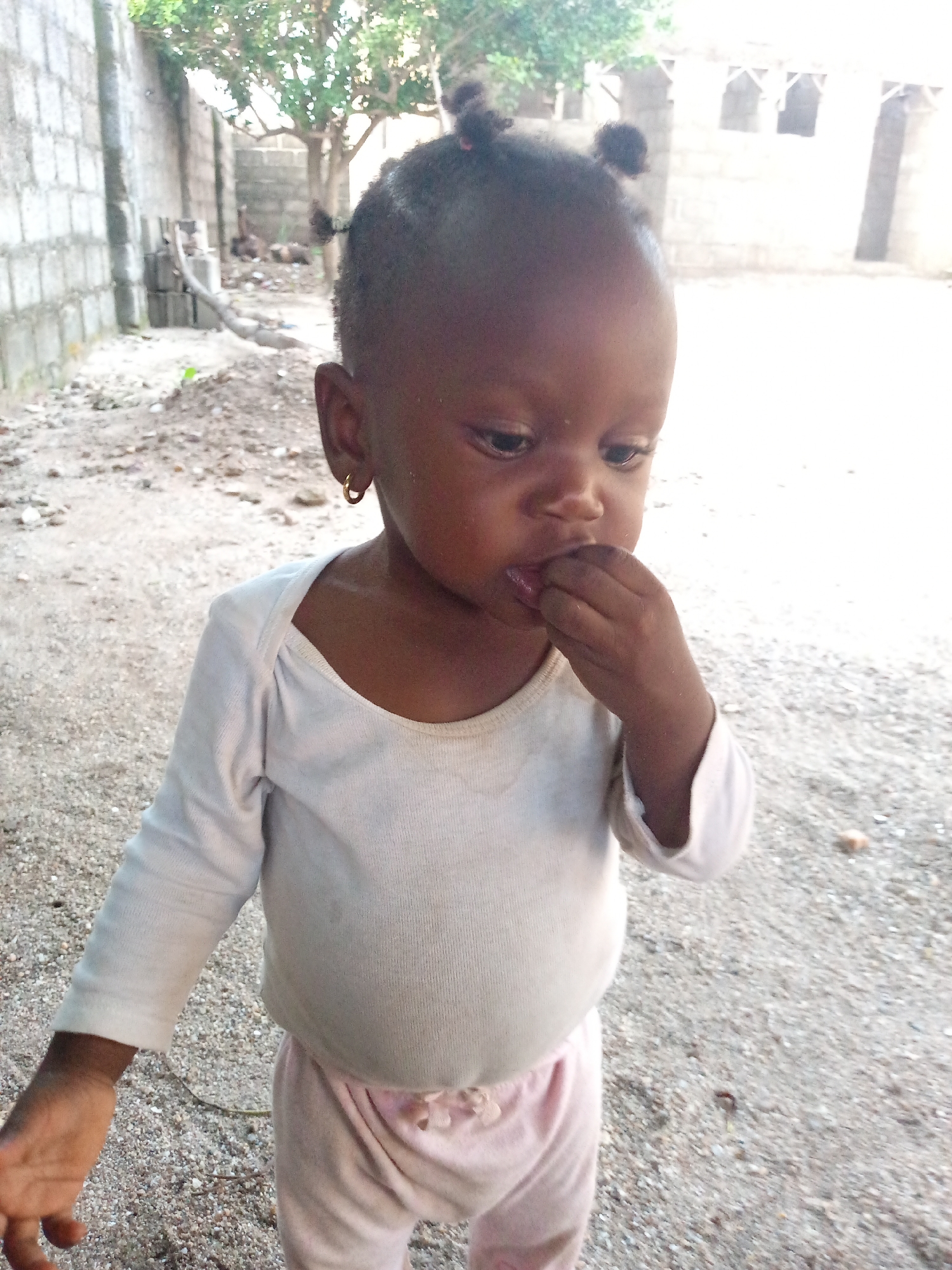 A young child with dark skin stands on a gravelly surface in an outdoor setting. The child wears a long-sleeved white shirt and light pink pants, and has small earrings. The child is looking down and has their fingers near their mouth. Behind the child, there are cinder block walls, a tree, and what appears to be the unfinished structure of a building. The location appears to be an impoverished area in an unknown city.