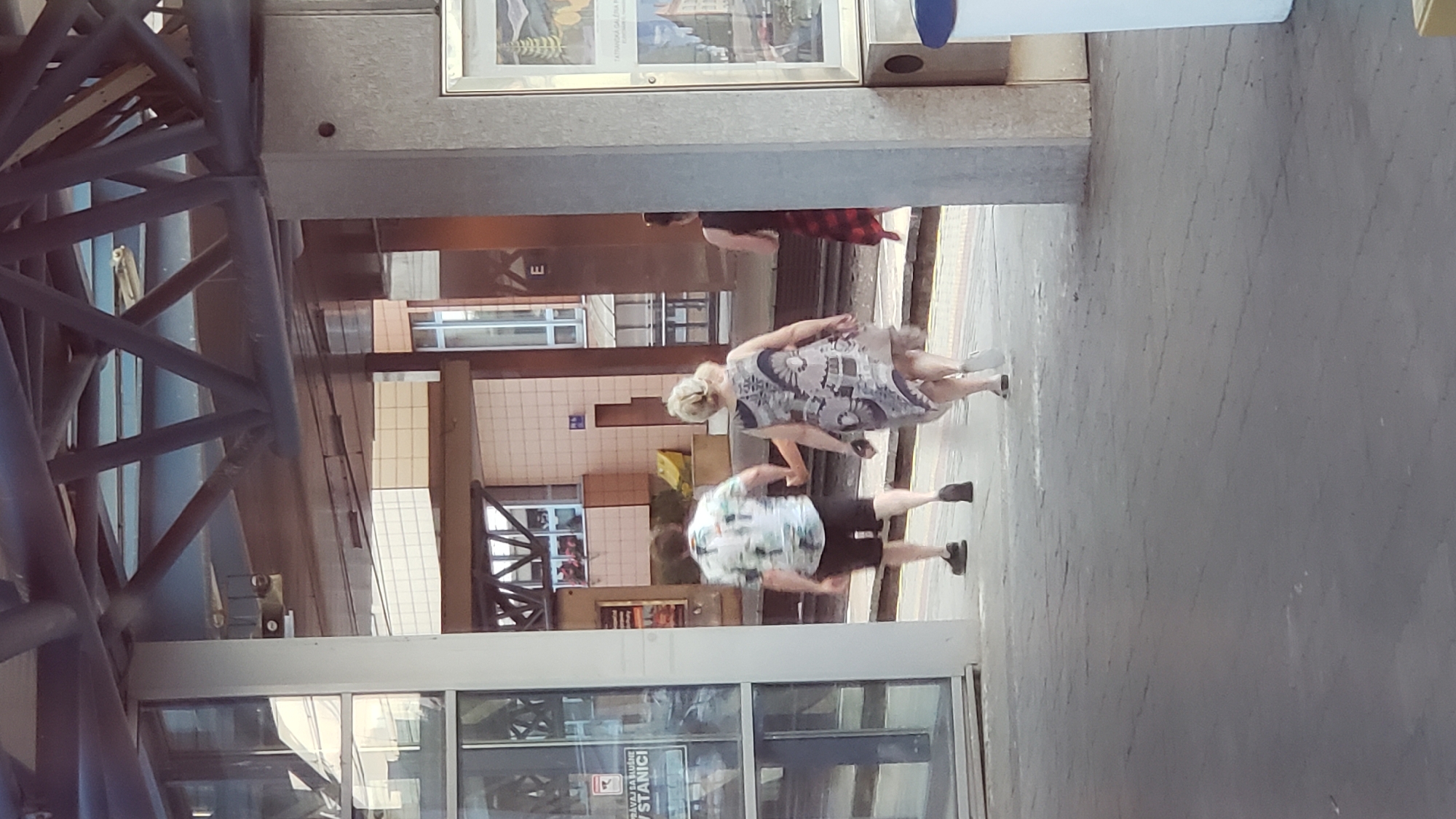 At the train station in Poprad, Slovakia, two individuals walk away from the camera. The person on the left, a male, is wearing a white shirt with a colorful pattern and dark shorts. The person on the right, a female, is wearing a patterned dress. They are walking on a paved platform next to train tracks. In the background, there are buildings with windows and architectural features typical of a train station. A third person, a female wearing a red and black plaid skirt, is visible further to the right, also walking away from the camera.