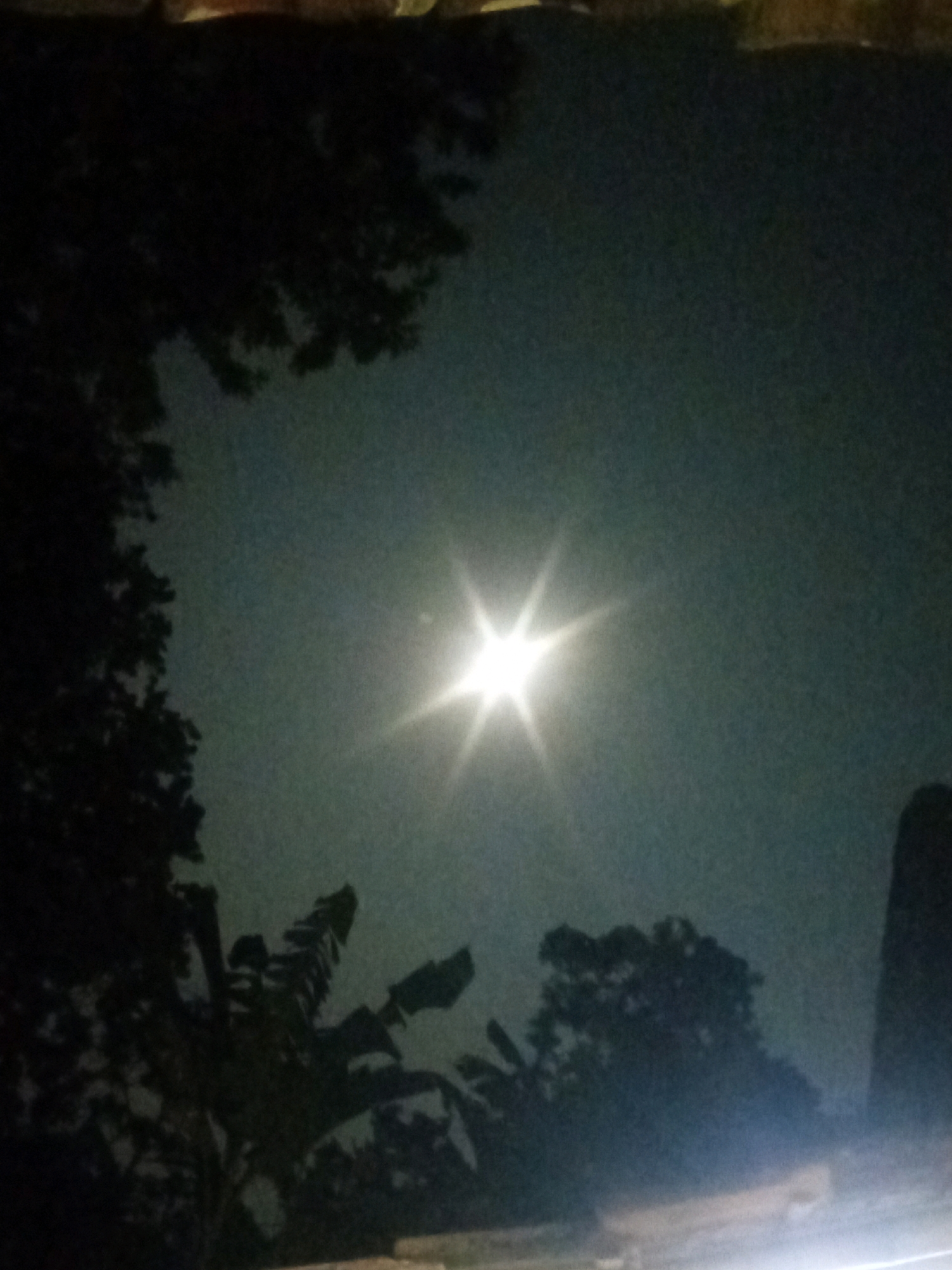 A nighttime outdoor scene depicts the moon as a bright, starburst-like object in a hazy sky. Silhouetted trees and foliage frame the lower and left portions of the image. The scene appears to be in Cempaka, Indonesia, based on the metadata. The foreground shows a blurry, indistinct surface, possibly a ground or rooftop, illuminated by an unseen light source.