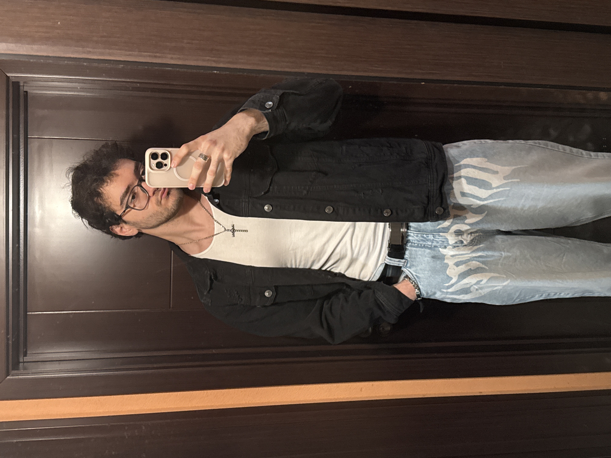 A young man is taking a selfie in front of a dark wooden door or possibly a mirror with a wooden frame. He has dark, curly hair, wears glasses, and has a light beard. He's wearing a white T-shirt, a black denim jacket, and light blue jeans with white flame-like patterns on the legs. He also wears a silver necklace with a cross pendant and a ring on his right hand. He's holding a smartphone, presumably taking the photo, with both hands. The location cannot be determined from the image, there is no indication this image was taken in Blagoevgrad, Bulgaria.