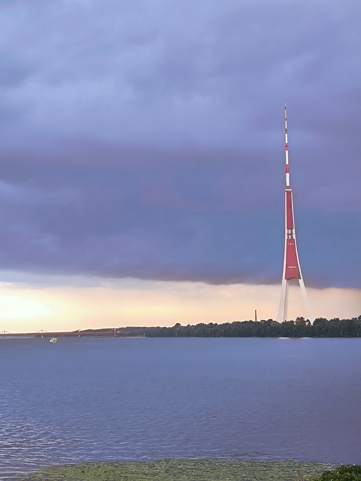 The image captures a wide shot of the Riga Radio and Television Tower, a prominent landmark in Riga, Latvia, situated on Zaķusala Island. The tower, a tall, slender structure with red and white segments, dominates the right side of the frame. Its distinctive lattice-work base is clearly visible, supporting the tapering spire. The tower is set against a dramatic sky filled with heavy, dark clouds in shades of blue and purple, suggesting an impending storm or the aftermath of one.
In the distance, across a body of water, a treeline forms a dark band along the horizon, punctuated by smaller industrial-looking structures. A bridge, likely part of the city's infrastructure, stretches across the water in the left mid-ground. A small white boat navigates the calm, blue water in front of the bridge, adding a sense of scale to the scene. The foreground of the image shows the surface of the water, rippled by a gentle breeze, with some aquatic vegetation visible at the bottom edge. The overall atmosphere is one of natural grandeur and the imposing presence of human engineering within a natural setting. There are no people visible in the image.