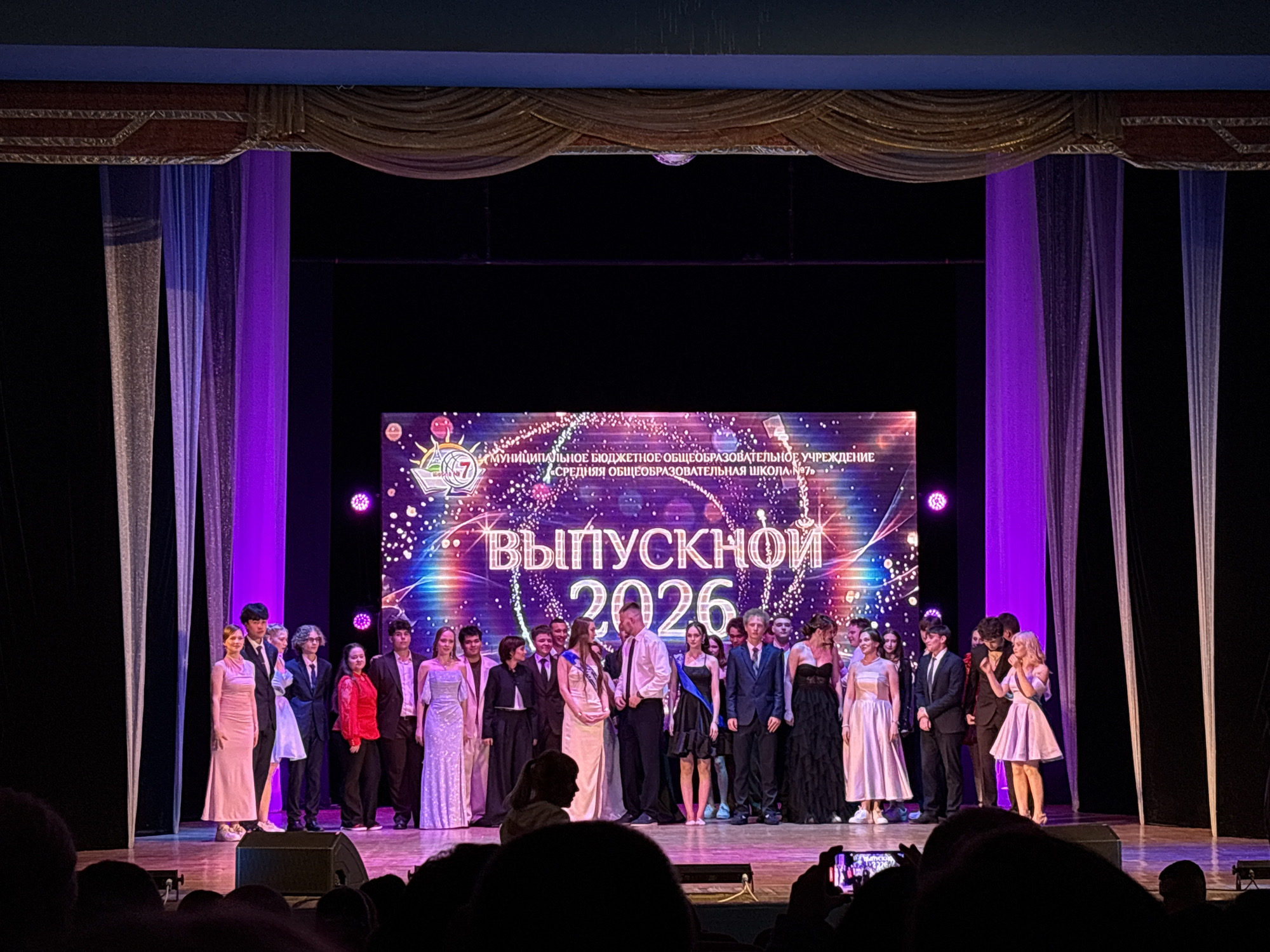 The image depicts a graduation ceremony on a stage, likely at a school in Birobidzhan, Russia, given the text on the screen. In the background, a large digital screen displays "ВЫПУСКНОЙ 2026" (Graduation 2026) in large white lettering, surrounded by colorful graphics and the name of an educational institution. The screen also shows text in Russian that translates to "MUNICIPAL BUDGETARY EDUCATIONAL INSTITUTION SECONDARY GENERAL EDUCATION SCHOOL No. 7".
On stage, a group of graduating students, dressed in formal attire, are gathered. Some are holding scrolls or wearing sashes. They appear to be posing for photographs or receiving recognition. The students are diverse in appearance, with both young men and women present. The lighting on stage is a mix of bright stage lights and purple uplighting from the side.
In the foreground, the audience is visible, with some individuals holding up their mobile phones to take pictures. The stage is set with curtains on the sides and a raised platform. The overall atmosphere suggests a celebratory and formal event.