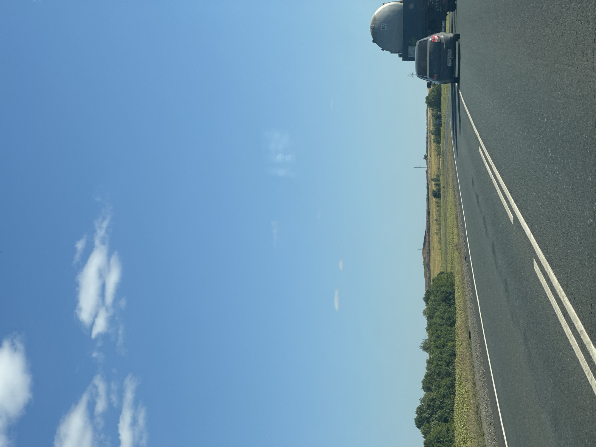 A dark-colored sedan is driving on a paved road under a bright blue sky with scattered white clouds. A large tanker truck with the designation "SV" is to the right of the sedan. The road is bordered by a grassy shoulder and then a field of green crops and trees. In the distance, there are rolling hills and more fields. The scene appears to be in a rural or semi-rural area. There are no people visible in the image.