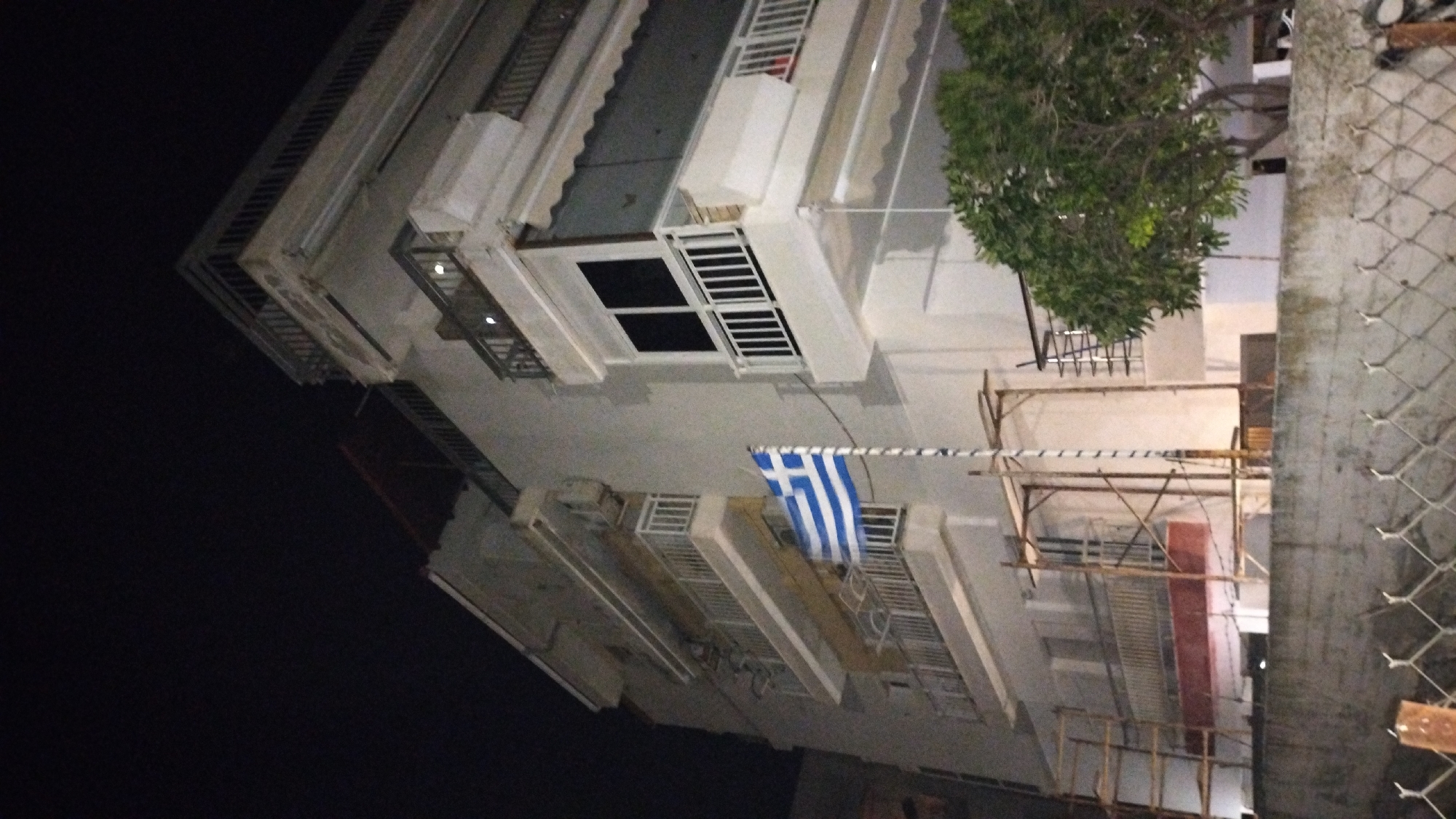 The image shows a multi-story building at night in Perea, Greece. A Greek flag is prominently displayed on a flagpole in front of the building, extending from approximately the second to the fourth floor. The building appears to be made of concrete and has several balconies with white railings and awnings. Scaffolding is visible on the lower levels of the building. A tree with green foliage is located to the right of the building, and a concrete wall with a wire fence runs along the bottom of the image. The scene is illuminated by artificial light, casting shadows and highlighting the textures of the building and its surroundings. The sky is dark, indicating nighttime.