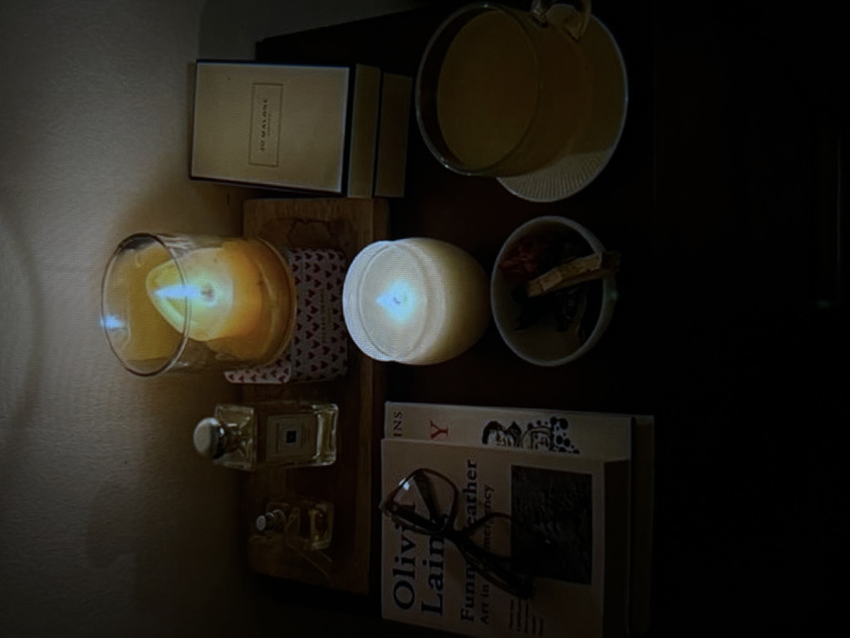 A dimly lit, warm-toned image displays a shelf with various items. A lit candle in a glass container sits alongside a box labeled "Jo Malone". A white candle is next to a small bowl filled with potpourri. A glass mug with a handle is visible. Below, two perfume bottles rest near a stack of books, one titled "Olivia Laing". A pair of glasses is atop the books. The image has a cozy, inviting feel.