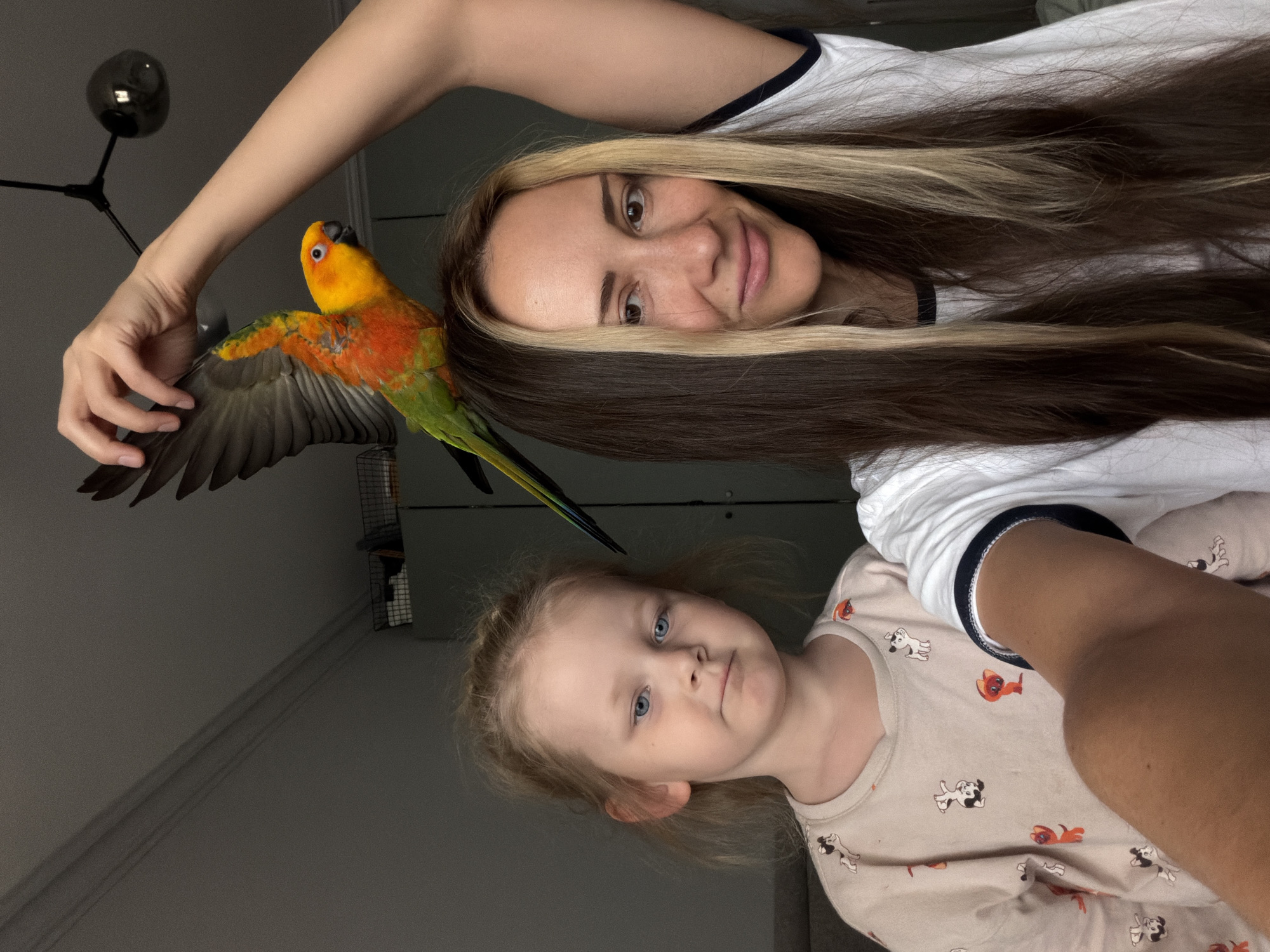 A selfie-style image shows a woman and a young child with a Sun Conure parrot in an indoor setting, seemingly a residential space.
The woman, positioned at the top of the frame, is fair-skinned with long, straight hair that is both blonde and brown. She is wearing a white t-shirt with a dark blue collar.
Below her, the young child has fair skin and blonde hair, and is wearing a pajamas with puppy figures.
The parrot is perched on the woman's hand with an open wing. The room appears dimly lit with gray walls. The corner of what appears to be a cage is visible in the background. A modern lamp is also seen.
There is nothing in the picture to indicate the location as Khimki, Russia.
