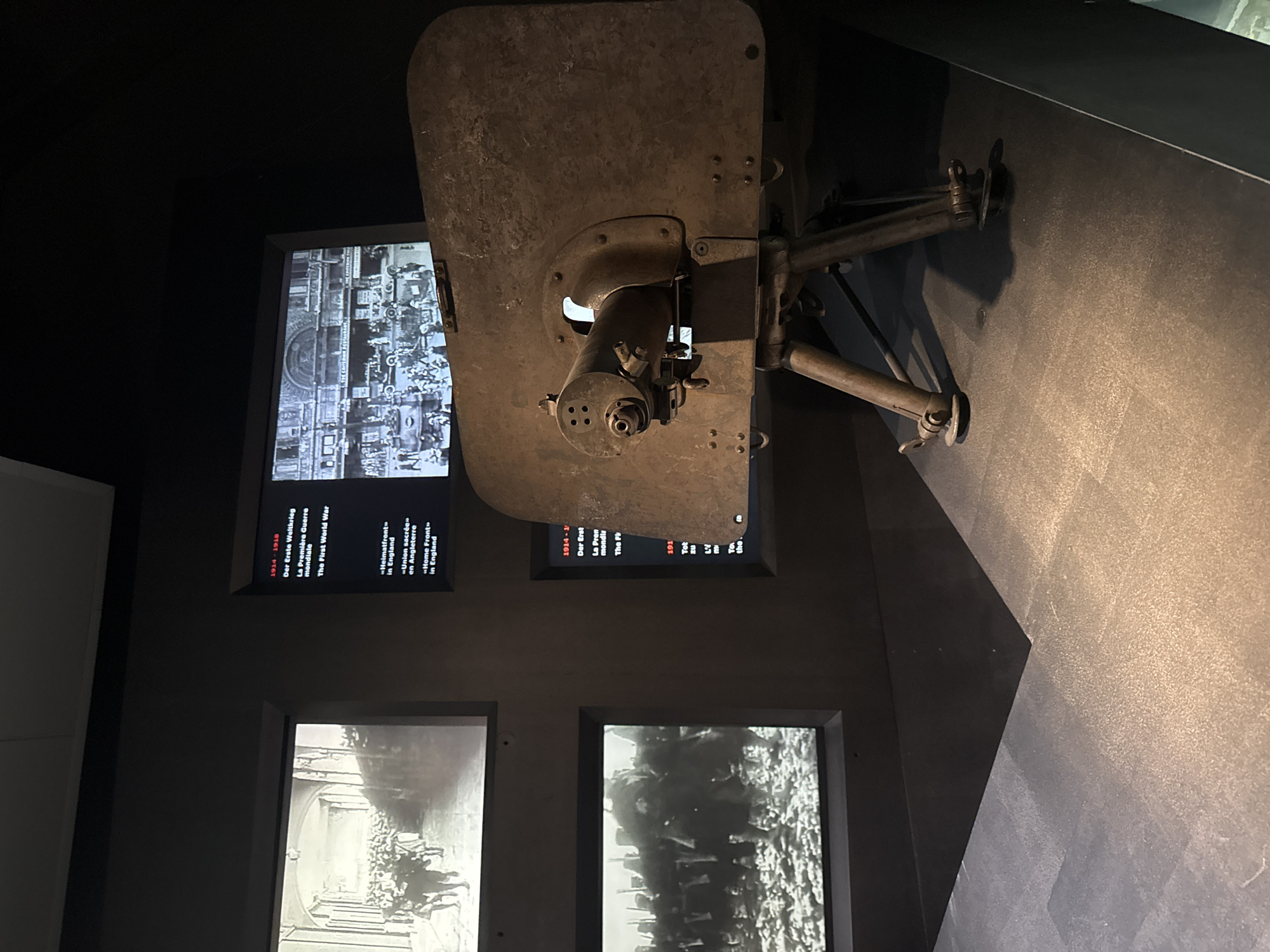 This media file depicts a museum exhibit related to World War I, likely located in Bern, Switzerland. The central focus is a large, weathered metal object, possibly a piece of military equipment or artillery, mounted on a stand. The object is positioned diagonally across the frame, obscuring parts of the background displays.
To the left of the equipment, two screens are visible. The upper screen displays a black and white historical photograph, likely depicting a scene from the war, with many people gathered. Below this, another screen shows text in multiple languages, including German, French, and English, with dates and titles such as "1914-1918," "Der Erste Weltkrieg," "La Première Guerre mondiale," and "The First World War." Further text indicates themes like "Heimatfront" and "Home Front."
Below these screens, two more screens are partially visible. The left one shows another black and white historical photograph, seemingly of people in procession or gathered in a formal setting with grand architecture. The right screen displays a black and white image of a large group of people, possibly soldiers or a crowd, in what appears to be an outdoor setting. The overall lighting is dim, typical of a museum environment, highlighting the displays. The setting appears to be an indoor exhibition hall. The images and text suggest a historical exhibition focusing on the impact of World War I.