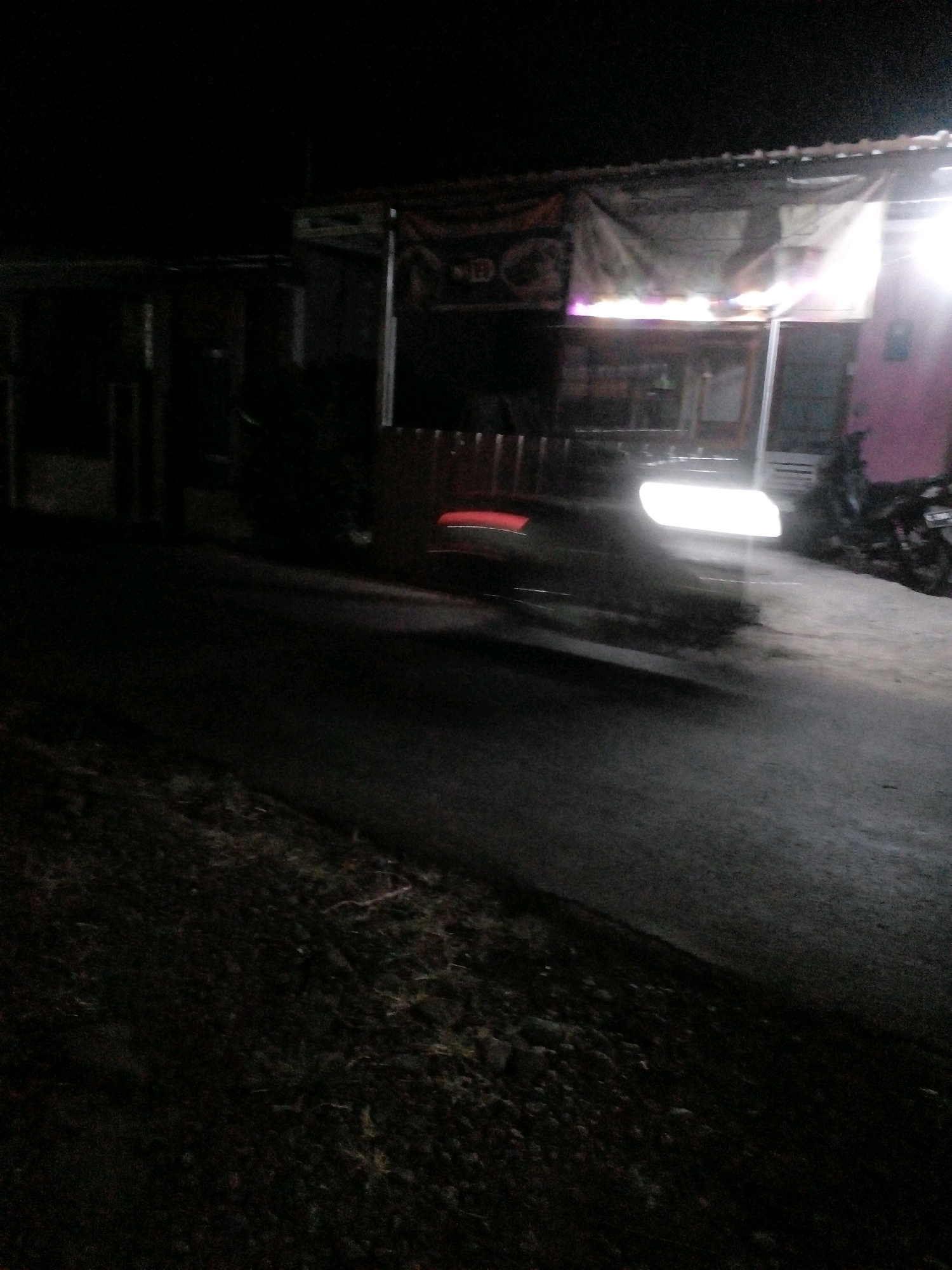 The image depicts a dimly lit street scene at night, likely in Pungangan, Indonesia, based on the provided context. In the background, a roadside stall with a canopy is visible, illuminated by bright lights. The stall appears to be selling food or goods, with some items visible on shelves or counters. A motorbike is parked to the right of the stall. The road in front of the stall is paved and appears to be wet, reflecting the lights. On the left side of the road, there is a grassy area with some debris. A vehicle's headlights are prominently displayed on the road, suggesting it is either stopped or moving. The overall impression is of a humble, functional roadside setup in a rural or semi-urban environment at night.