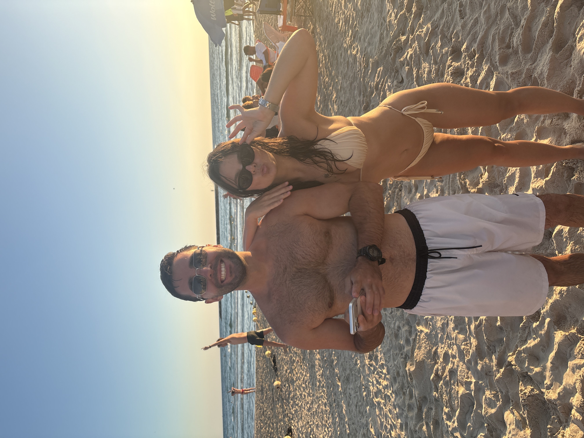 This is a candid, eye-level, full-body shot taken at sunset on a sandy beach in Tel Aviv, Israel. A couple is the primary focus, positioned centrally in the frame. The male subject, on the left, is shirtless and wearing white swim shorts. He has short, dark hair, a beard, and is wearing sunglasses. He is holding a smartphone in his right hand and looking towards the camera. The female subject, on the right, is wearing a beige bikini. She has long, dark, wet hair and is also wearing sunglasses. She is puckering her lips and holding her right hand up to her face in a playful gesture, looking at the camera. Her left arm is resting on the male subject's shoulder.
The background shows a beach scene with the Mediterranean Sea in the distance. The water is calm, and there are other beachgoers visible. To the left, further down the beach, two people are standing in the shallow water. To the right, an umbrella with the logo "Wolt" is visible, along with some beach chairs and tables. The sand is light brown and textured, with footprints and shadows from the low-angled sunlight. The sky is a clear gradient of orange and blue, indicating the sun is setting. The overall atmosphere is relaxed and summery.