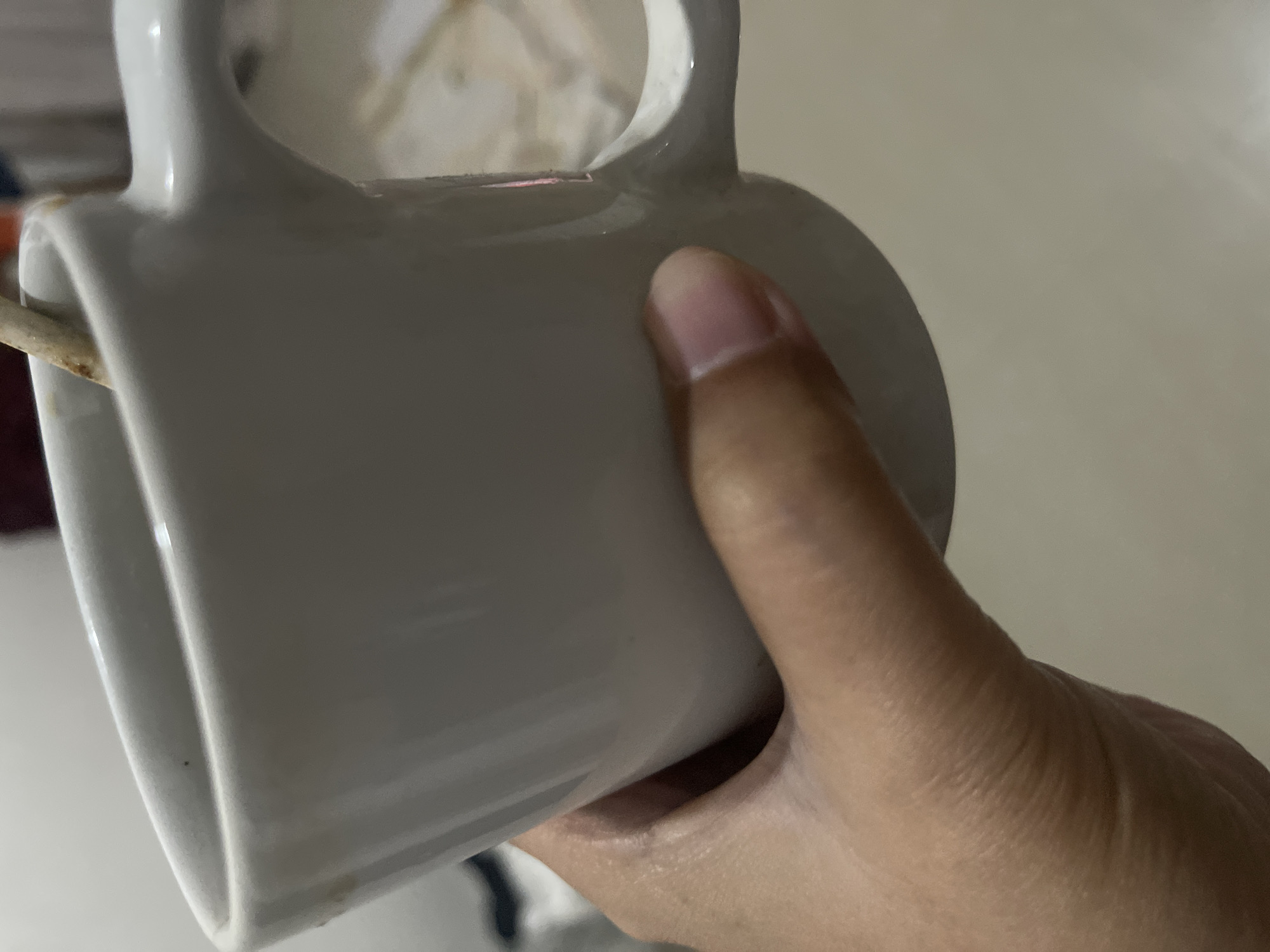 A person's hand is holding a white ceramic mug. The mug appears to be empty, and its handle is partially obscured by the hand. A wooden skewer or stick is visible, inserted through the handle of the mug. The background is blurred and out of focus, suggesting an indoor setting. The lighting is neutral, not indicating a specific time of day or weather conditions. No text is visible in the image. The focus is on the interaction between the hand and the mug.