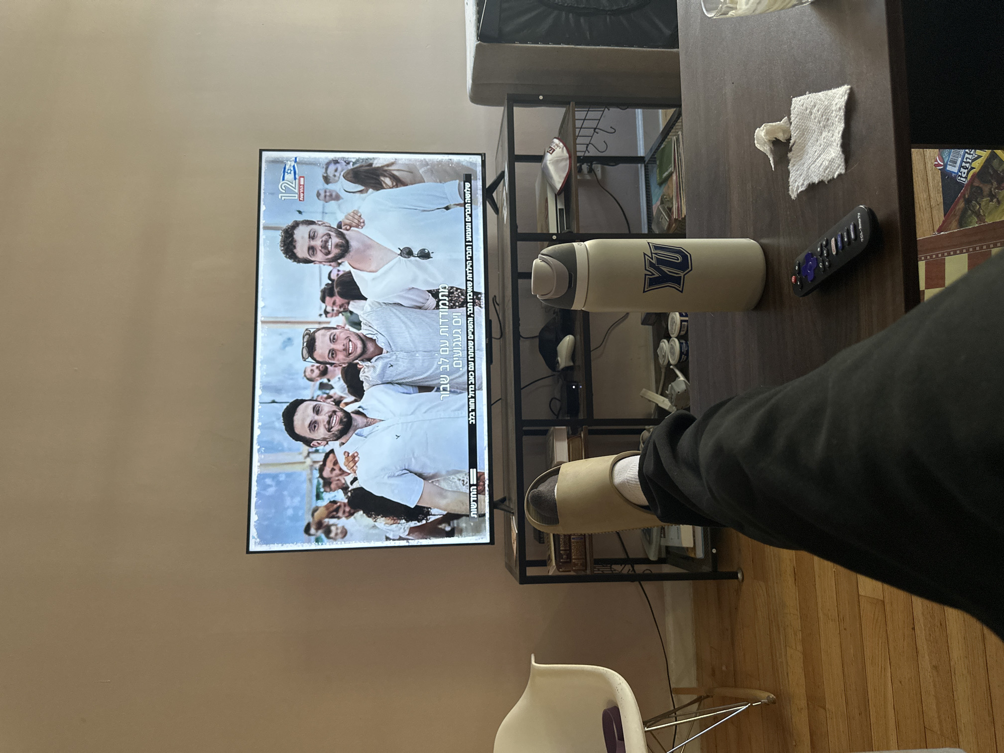 The image captures an indoor scene, likely a living room or den, in New York, USA, from the perspective of someone relaxing. In the foreground, a person's lower leg and foot, dressed in black pants, white socks, and a light-colored slipper, are visible. The foot extends over a dark brown wooden coffee table.
On the table, a beige water bottle with a blue "YU" logo stands next to a black Roku TV remote control and a piece of crumpled white paper towel. Behind the table, a black metal shelving unit is visible, holding various items, including a white and red kippah or yarmulke on one of the shelves.
To the left, a flat-screen television displays what appears to be a news program. The screen shows a group of smiling men, with three prominent figures wearing white shirts. Visible Hebrew text on the screen includes "12" and "החדשות" (The News). Below the main image, additional Hebrew text reads: "מתמודדות עם לב שבור וים געגועים. שלושה חברים וגעגוע | חברי הילדות שאיבדו חבר, והשניים שנותרו עם כאב גדול וחור בלב," which translates to "Coping with a broken heart and much longing. Three friends and longing | childhood friends who lost a friend, and the two who remained with great pain and a hole in the heart." This text suggests a segment about loss or grief, despite the smiling faces on screen. The floor is covered with light-colored wooden planks. A white chair is partially visible in the lower left corner.