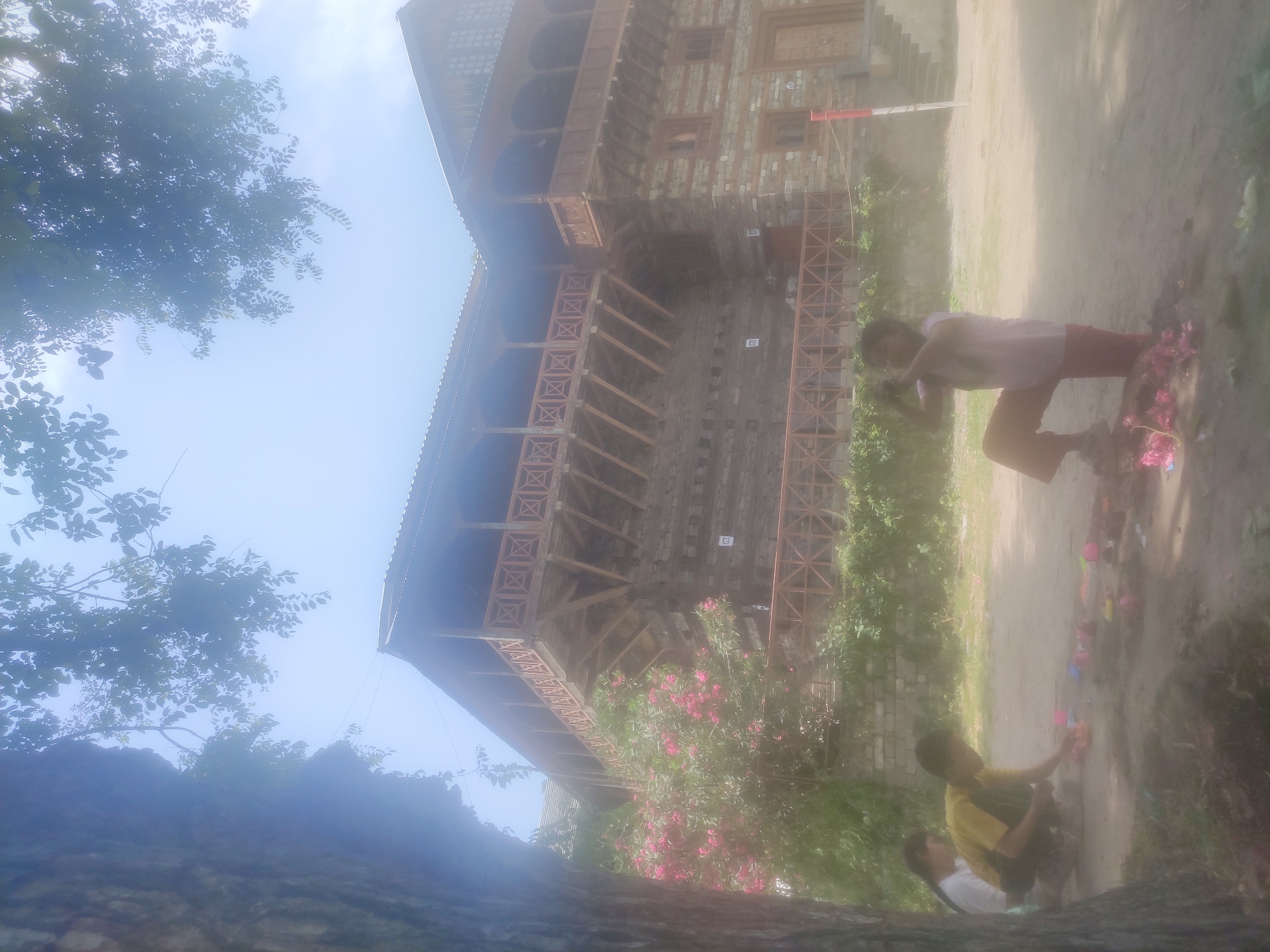 In Naggar, India, a weathered stone and wood building with traditional balconies stands under a bright sky, partially obscured by foliage. In the foreground, two children are playing on a dirt ground. A boy in a yellow shirt is kneeling, arranging colorful toys. A girl in a pink shirt and red pants is standing, also near toys, with a pink flower at her feet. A third child, partially visible, is seated to the left, wearing white. The scene is hazy, suggesting either dust or a soft focus effect.