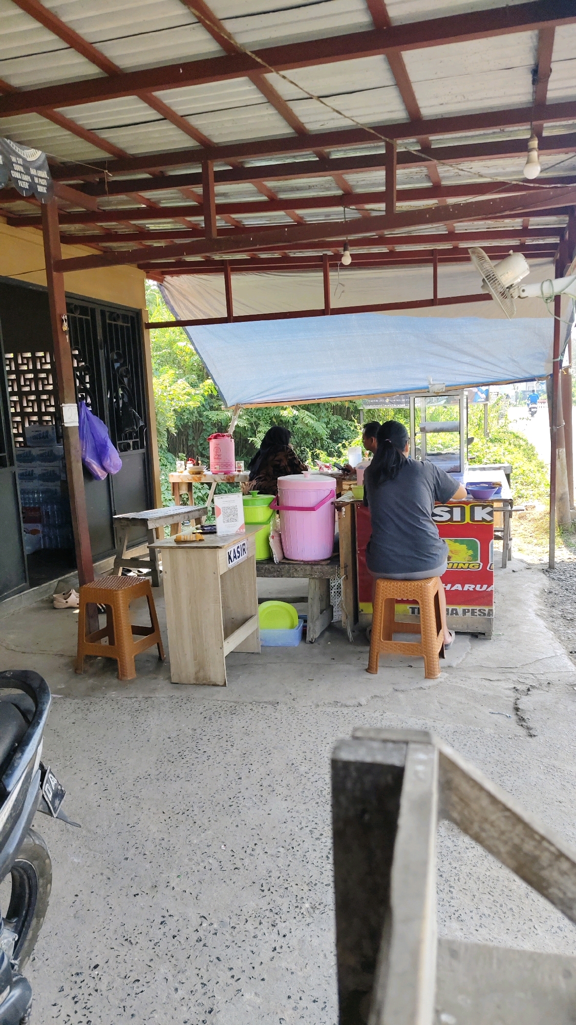 The image depicts an outdoor food stall operating under a corrugated metal roof supported by wooden beams. The stall is situated on a concrete floor, likely in Banjarmasin, Indonesia, suggested by the context. Two individuals are visible, one appears to be a cashier at a small wooden counter labeled "KASIR" (cashier), and another person, possibly a customer, is seated at the food counter. The food stall is equipped with containers of pink and green buckets, a water dispenser, and various other items associated with food preparation and service. Behind the stall, lush green foliage indicates a natural environment. In the foreground, a motorcycle is partially visible, suggesting it is parked nearby. The overall scene conveys a casual, everyday setting of a local food establishment in a tropical region.