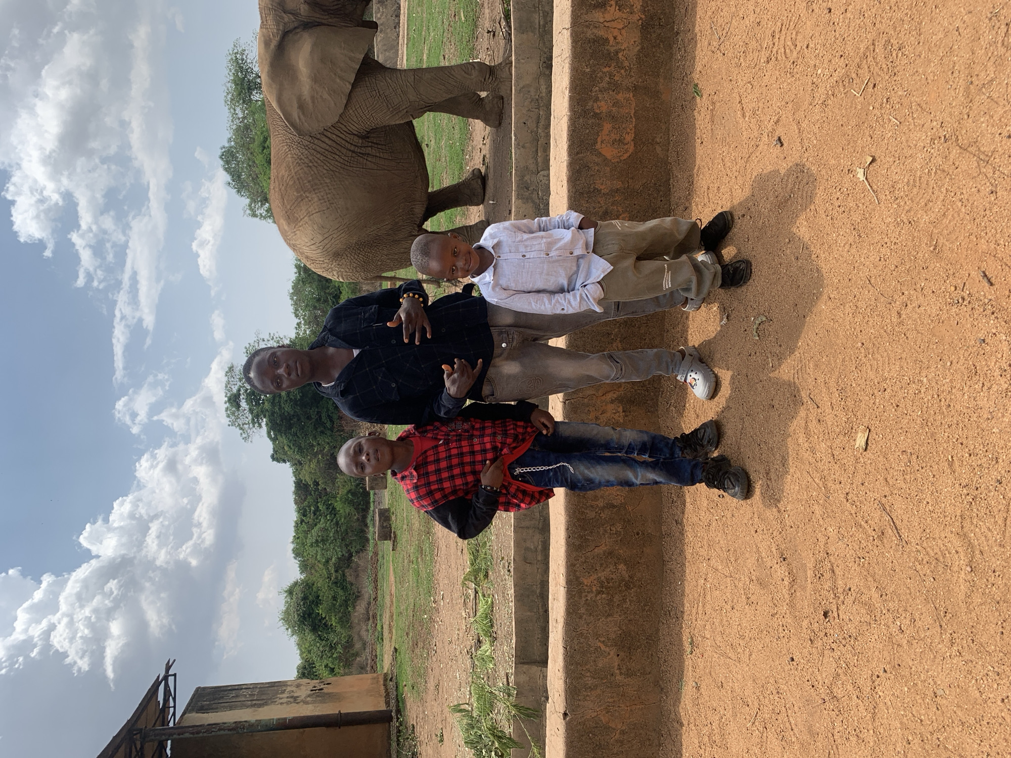 A family of three poses for a photograph near an elephant enclosure at Jos Wildlife Park in Jos, Nigeria. The image is taken during daylight hours with a partially cloudy sky. A man, presumably the father, stands between his two sons. The older son wears a red and black flannel shirt with jeans and black shoes. The younger son wears a white shirt with tan pants and grey and white shoes. The father is wearing a black shirt, grey pants, and white shoes. The elephant enclosure is visible in the background, with a partial view of the elephant. The environment includes green foliage in the background, a red-brown dirt ground, and concrete barriers. A partial structure is visible in the lower left corner.