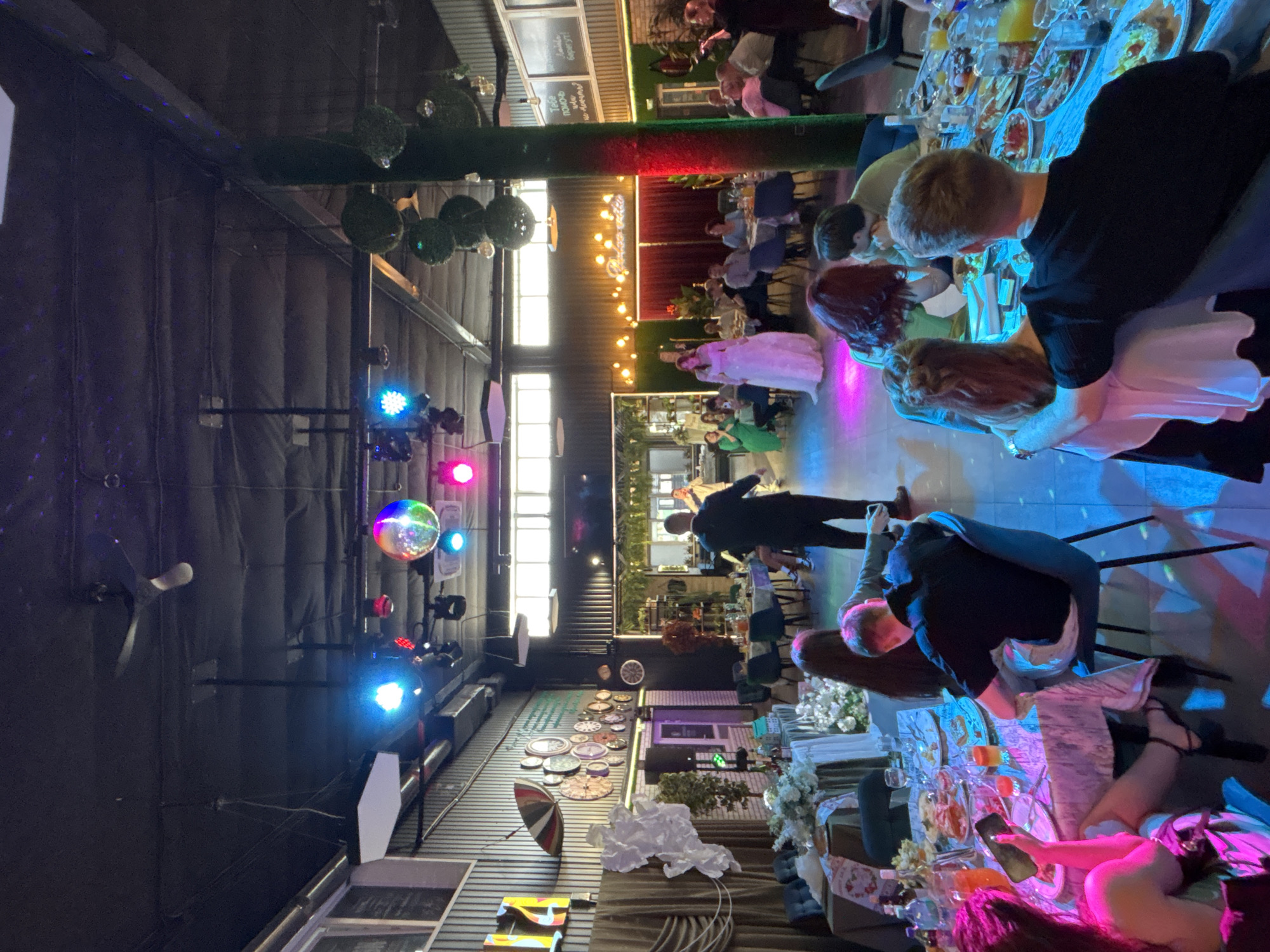 The image depicts a festive indoor gathering, possibly a wedding reception or a party, held in Chernushka, Russia. The scene is illuminated by a variety of colorful lights, including blue, pink, and disco ball effects, creating a vibrant atmosphere. Several round tables are set up, adorned with white tablecloths, floral arrangements, and food platters, indicating a meal is in progress.
In the foreground, several individuals are seated at a table, engaged in conversation and looking at their phones. One person is holding a smartphone, illuminated by its screen. In the middle ground, a man in a suit is standing, seemingly addressing a group of people. A woman in a long white dress, likely the bride, is positioned near the center of the room, facing away from the camera. A line of guests are seated at tables in the background, observing the events.
The environment appears to be a banquet hall or a restaurant with industrial-style walls and exposed ceilings. Decorative elements include a large green column covered in artificial foliage and a wall adorned with numerous clocks. The overall impression is one of a lively and celebratory occasion.