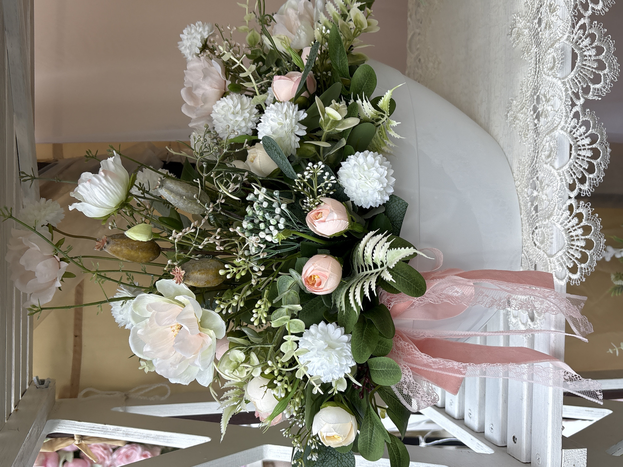 The image displays a floral arrangement in a white oval vase, adorned with pink ribbons and lace. The arrangement features a variety of artificial flowers and greenery, including large pale pink peonies, white chrysanthemum-like blooms, small pink rosebuds, and delicate white filler flowers, interspersed with green leaves and dried seed pods. The vase sits on a white tiered shelf, which appears to be part of a decorative display or furniture item, potentially a shelf unit or a stand. The background is a soft, pale pink wall, and a white lace doily or tablecloth is visible beneath the vase. The lighting suggests an indoor setting with diffused natural light. The arrangement is predominantly in pastel shades of pink, white, and green. The scene does not contain any people or any identifiable landmarks or details that would suggest a specific location in the city of Vitsebsk, Belarus.