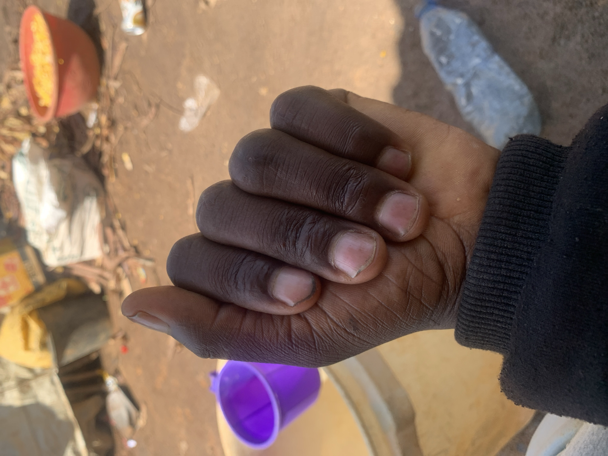A close-up, first-person perspective shot shows a clenched fist with dark skin and trimmed fingernails, partially obscuring the background. The hand is covered by the sleeve of a black sweatshirt. In the blurred background, a purple plastic cup sits within a larger light-colored bucket. Further back, a red bowl filled with yellow kernels, possibly corn, is visible, alongside a clear plastic water bottle lying on the ground. The ground appears to be dirt with scattered debris. The scene suggests an outdoor environment, possibly in Buga da Gindi, Nigeria.