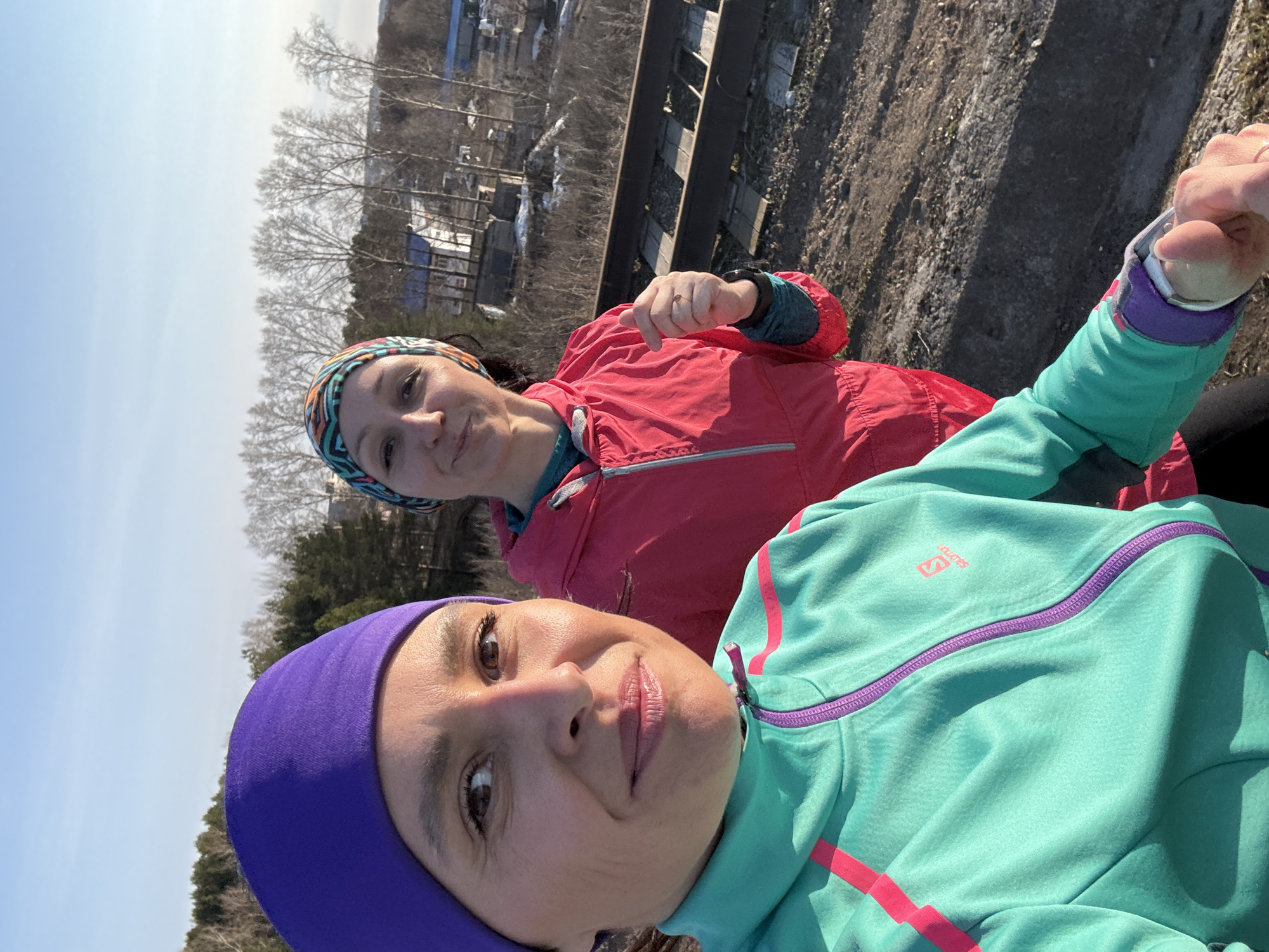Two women pose for a selfie in Chaikovsky, Russia, near railway tracks. One woman wears a purple head covering and a turquoise jacket with a purple zipper and pink stripes. The other woman wears a colorful headband and a red jacket. In the background are buildings, trees, and a blue sky.