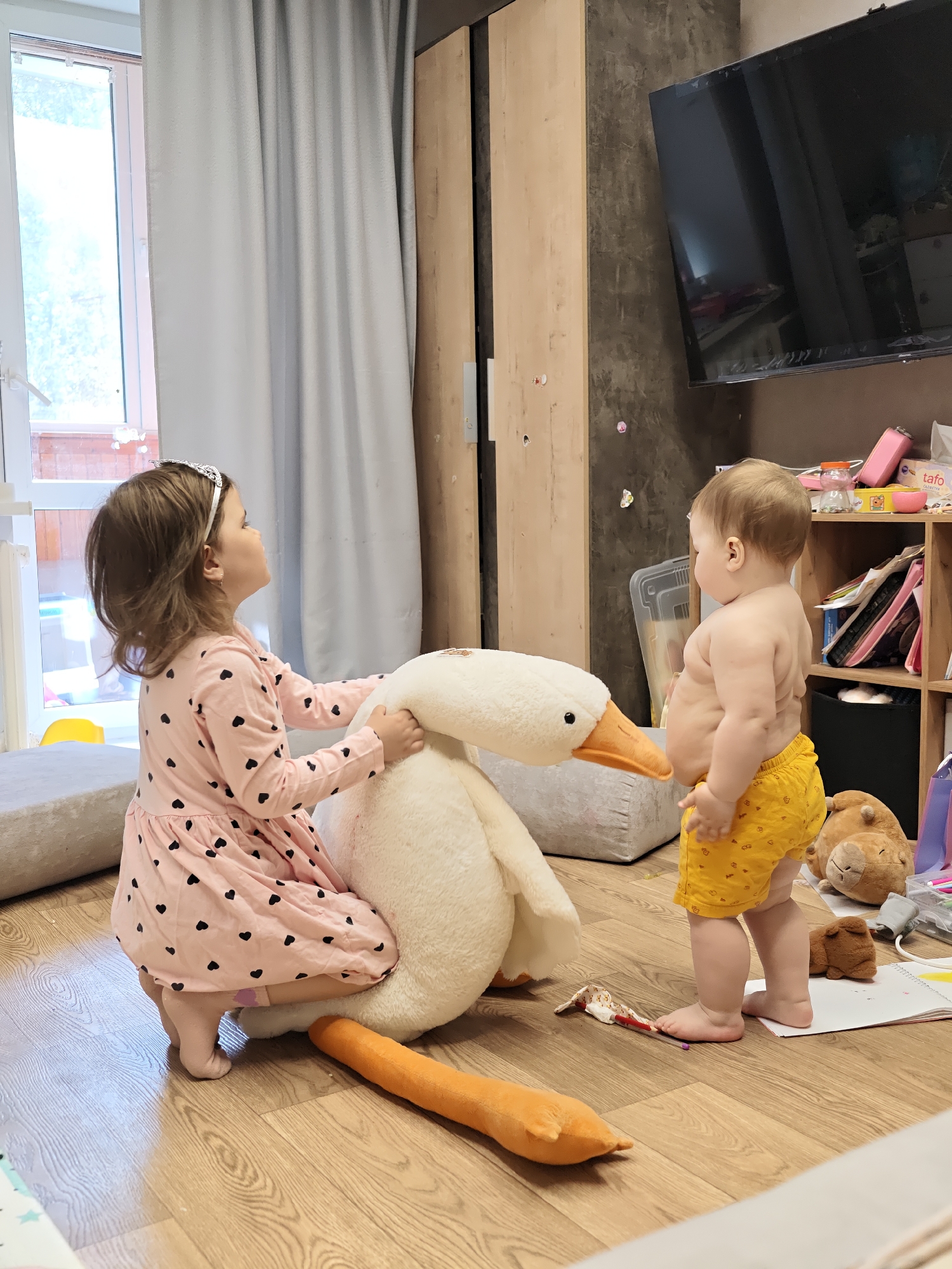 The image shows an indoor scene with two young children in a room with light wood-grain flooring. A girl, wearing a light pink dress with black heart patterns and a silver tiara, is kneeling next to a large, stuffed white goose. She is looking up to her left, and her hands are touching the goose's head. Beside her, a baby, wearing only yellow shorts, is standing and facing towards the goose. The baby has a round belly and the chubby texture of a baby's skin. Between the children and in front of the goose is a stuffed orange carrot toy and a torn piece of fabric. Behind the baby is a shelf unit filled with various items, including containers, books, and stuffed toys. A large television is mounted on the wall above the shelf, and to the left is a tall wooden cabinet and a curtained window. Pillows are visible in the background. The scene appears to be from a domestic setting, possibly a child's playroom or living room. The presence of both curtains and a window suggests a room with natural light. The context of Chaikovsky, Russia is not discernible from this image.
