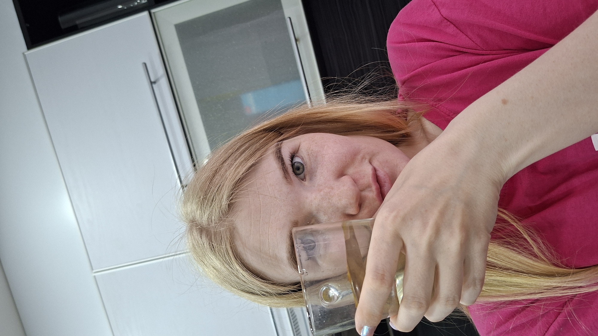 The image shows a blonde woman wearing a pink shirt, holding a glass with a clear liquid. She appears to be in a kitchen with white cabinets. The location is Chaikovsky, Russia.