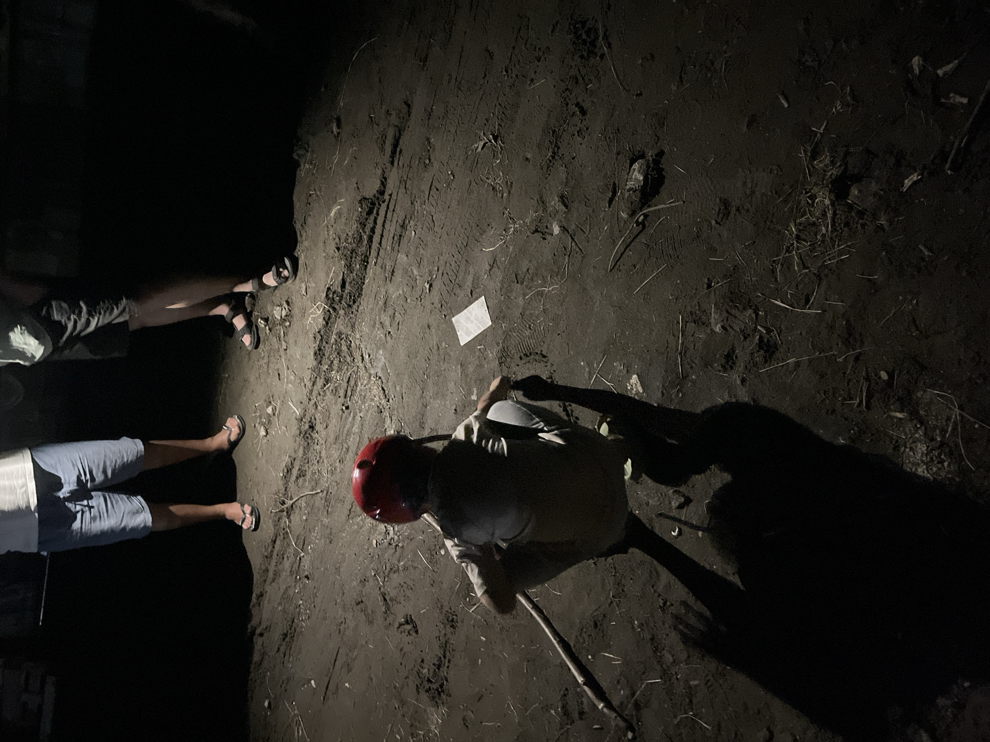 In the city of Poti, Georgia, a person wearing a red helmet and a light-colored shirt squats on dark, muddy ground at night. They are holding a long, thin stick with their left hand. To the right, a small white object lies on the ground. In the background, the legs of two other individuals are visible. One person wears light blue shorts and flip-flops, while the other wears dark shorts and sandals. The ground shows tire tracks and scattered debris. The lighting suggests artificial illumination, casting long shadows.