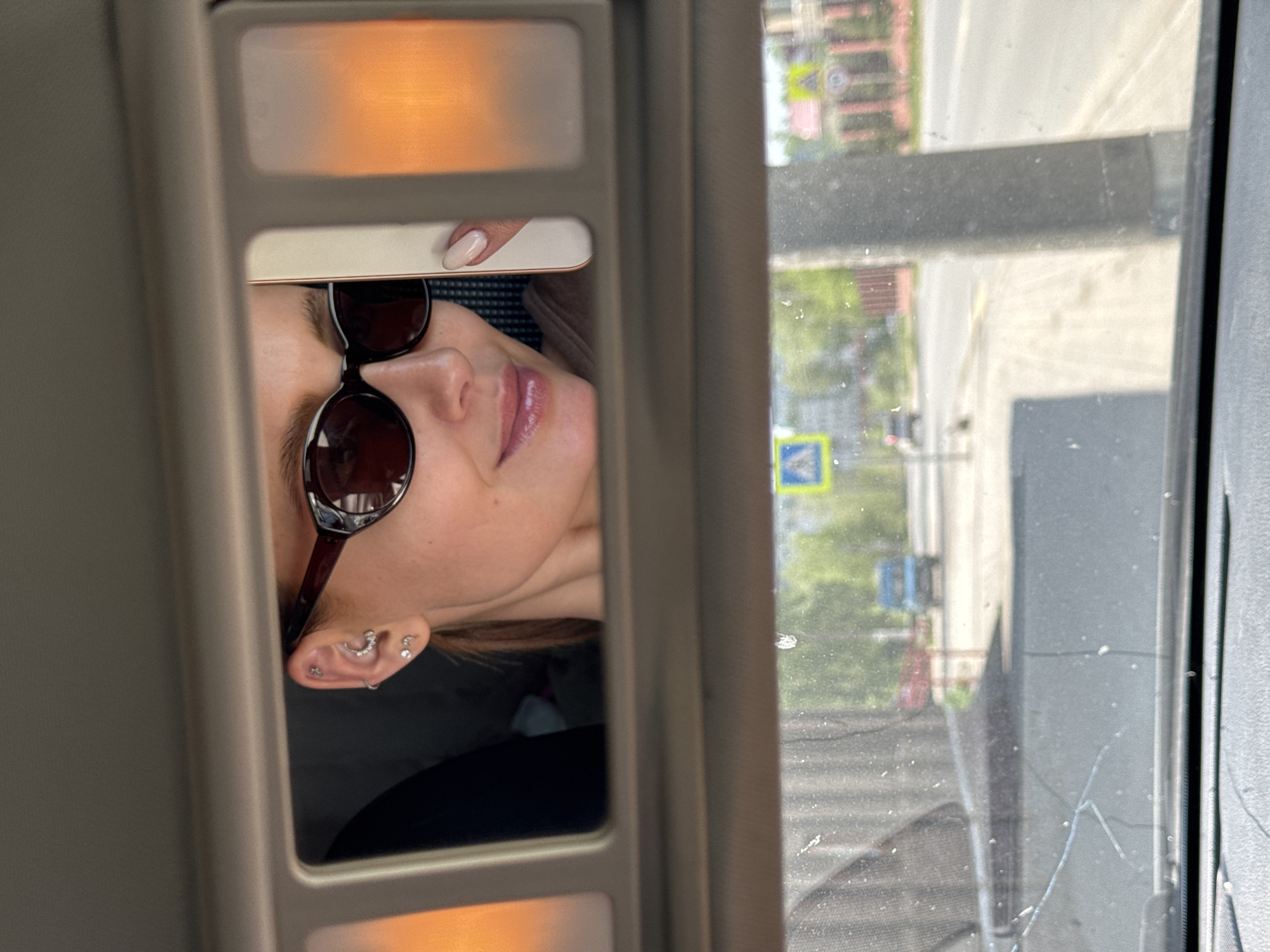 The image shows a view through a car's sun visor mirror and the adjacent side window. A woman's face, reflected in the mirror, is visible. She is wearing sunglasses and has multiple piercings in her left ear. Her right hand is near the top of the mirror. The mirror has two light panels, one at the top and one at the bottom, which appear to be illuminated. Through the car window, a street scene is visible, including buildings, trees, sidewalks, and a road. A pedestrian crossing sign is visible on the street. The window appears to be dirty and has a crack. The street sign suggests this photo was taken in Chaikovsky, Russia.