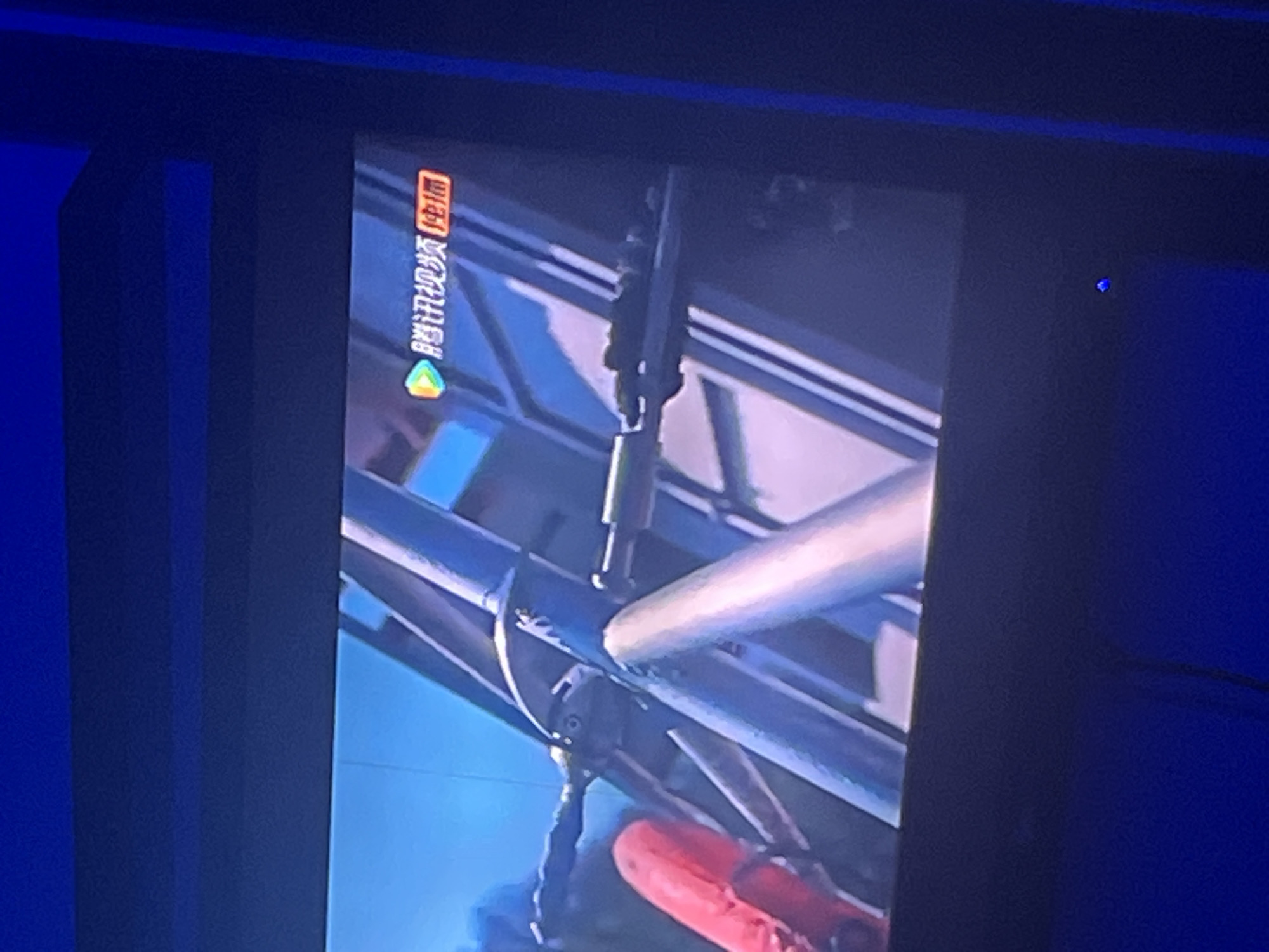The image displays a close-up, slightly blurry view of a television screen, likely in a dimly lit room. The screen shows a program with a logo in the upper right corner that reads "腾讯视频" and "企鹅" in Chinese characters, indicating it's a broadcast from Tencent Video. The visual content on the screen appears to be from a scene involving industrial or structural elements, possibly a construction site or a mechanical apparatus, with prominent blue and silver metallic beams. A red object is partially visible on the left side of the frame. The surrounding environment of the room is dark, with blue lighting accents visible, possibly from ambient light or fixtures. There is no discernible indication of the city of Jauro Yinu, Nigeria, within the image itself.