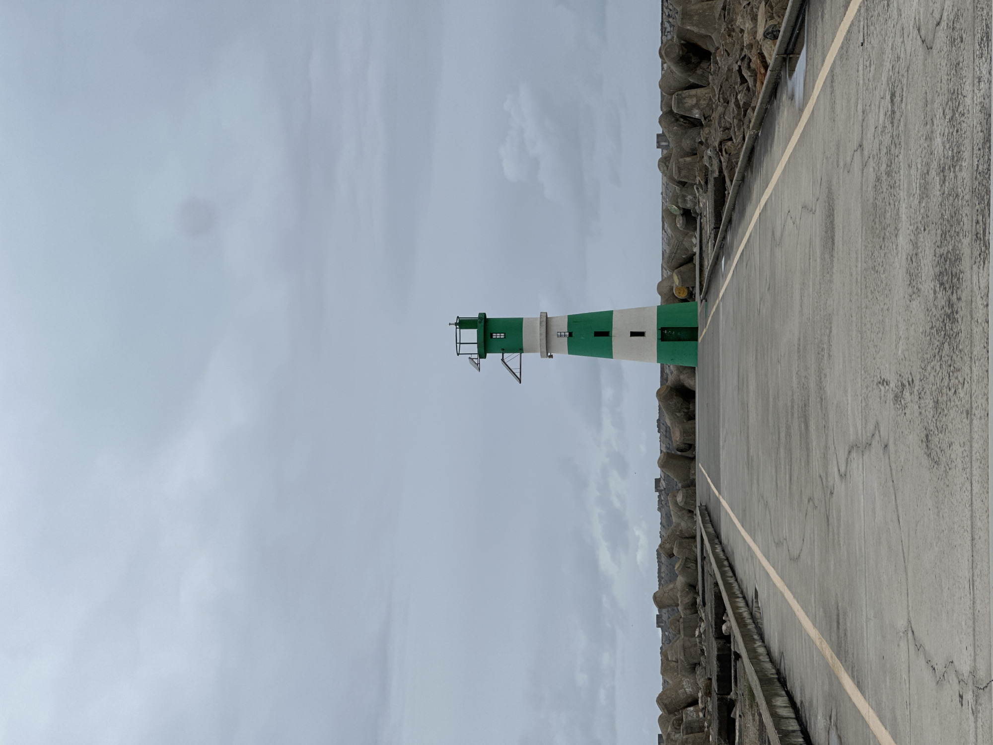 The image shows the Barra lighthouse in Gafanha da Nazaré, Portugal. It's a gray, overcast day. The lighthouse is white and green striped, situated next to a concrete pier with a yellow line marking the edge. A breakwater made of irregularly shaped concrete blocks runs along the side of the pier.